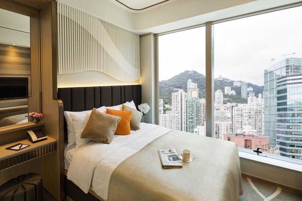 A modern hotel room with a large bed, a bedside table with a lamp, a mirror, and a view of the cityscape.