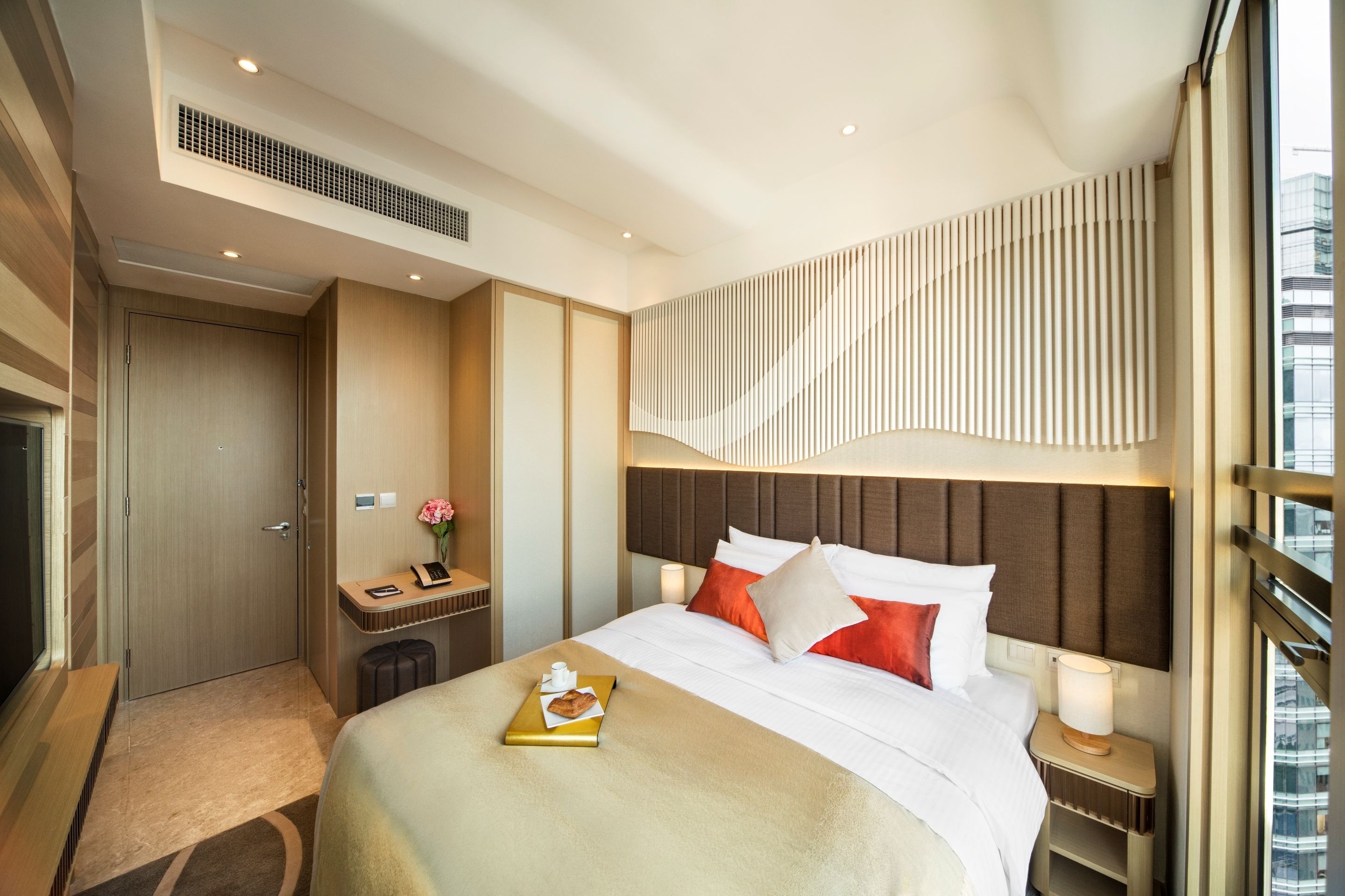 A modern hotel room with a large bed, a bedside table with a lamp, a small desk with a chair, and a large window offering a city view.