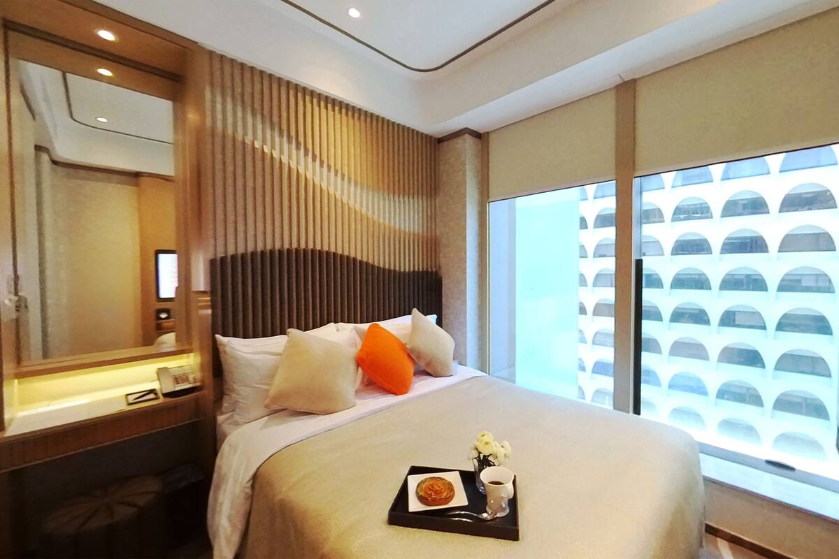 A hotel room with a large bed, a bedside table, a mirror, and a window with a view of a building.