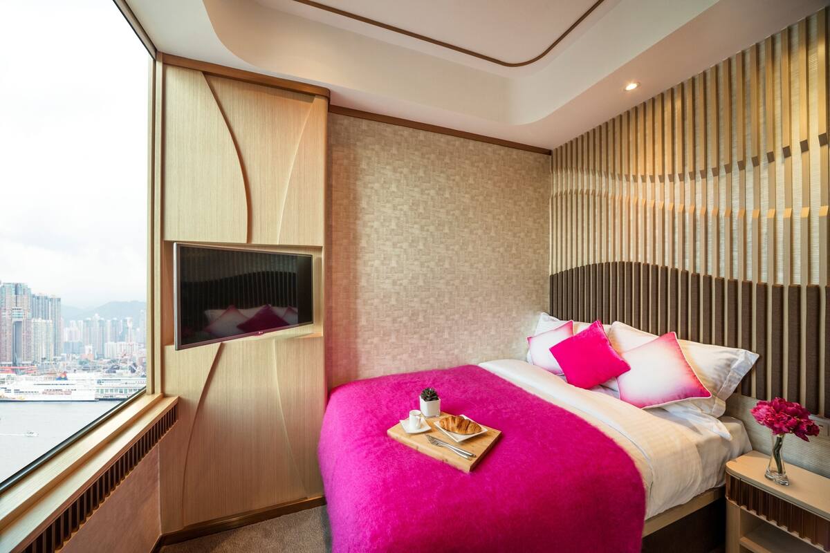A modern hotel room with a large bed, a flat-screen TV, a city view, and a wooden panel wall.