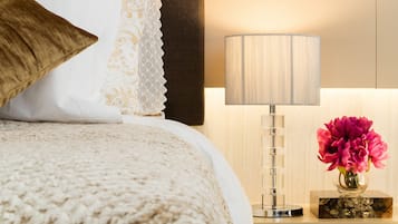Un lit bien fait, agrémenté d’un coussin blanc et doré à motifs, d’un coussin marron et d’une couverture texturée. On aperçoit une table de chevet avec une lampe et un vase de fleurs.
