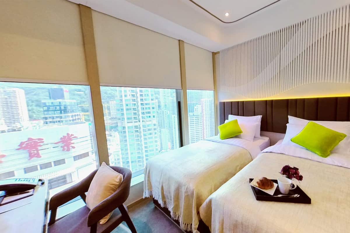 A hotel room with two beds, a desk with a chair, and a large window offering a city view.