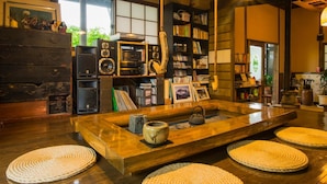 A cozy room with a wooden coffee table, woven mats, and a bookshelf filled with books and decorative items. - Yurari Rokumyo (Yufu)