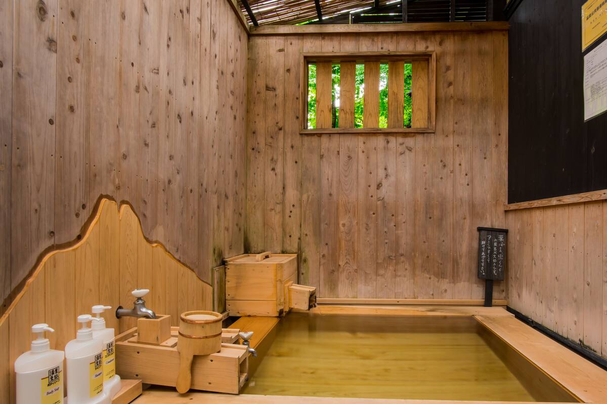 A wooden interior with a wooden bathtub, wooden benches, and a window with a view of trees.