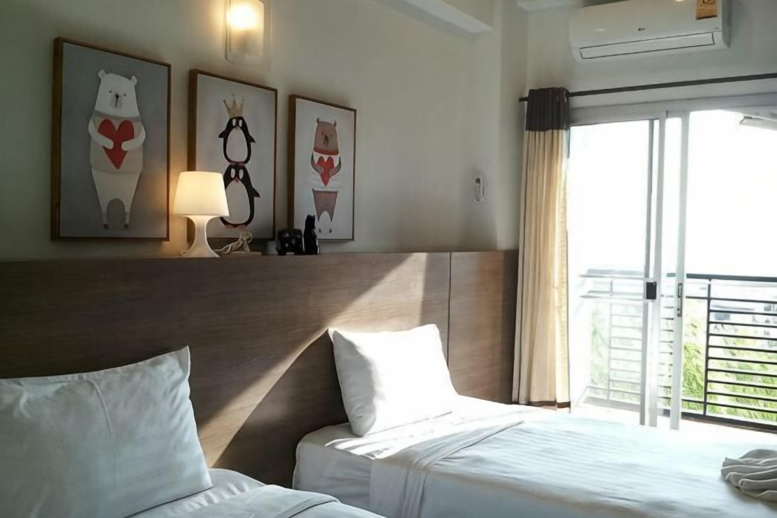A hotel room with two beds, a headboard, artwork on the wall, a lamp, and a view of the outdoors.