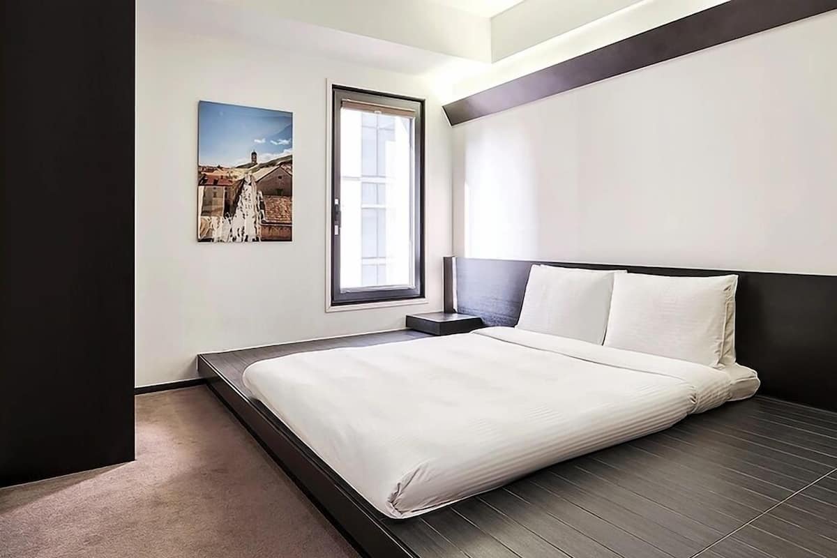 Ein modernes Hotelzimmer mit einem großen Bett, einem Nachttisch, einem Fenster mit Blick und einem gerahmten Bild an der Wand.