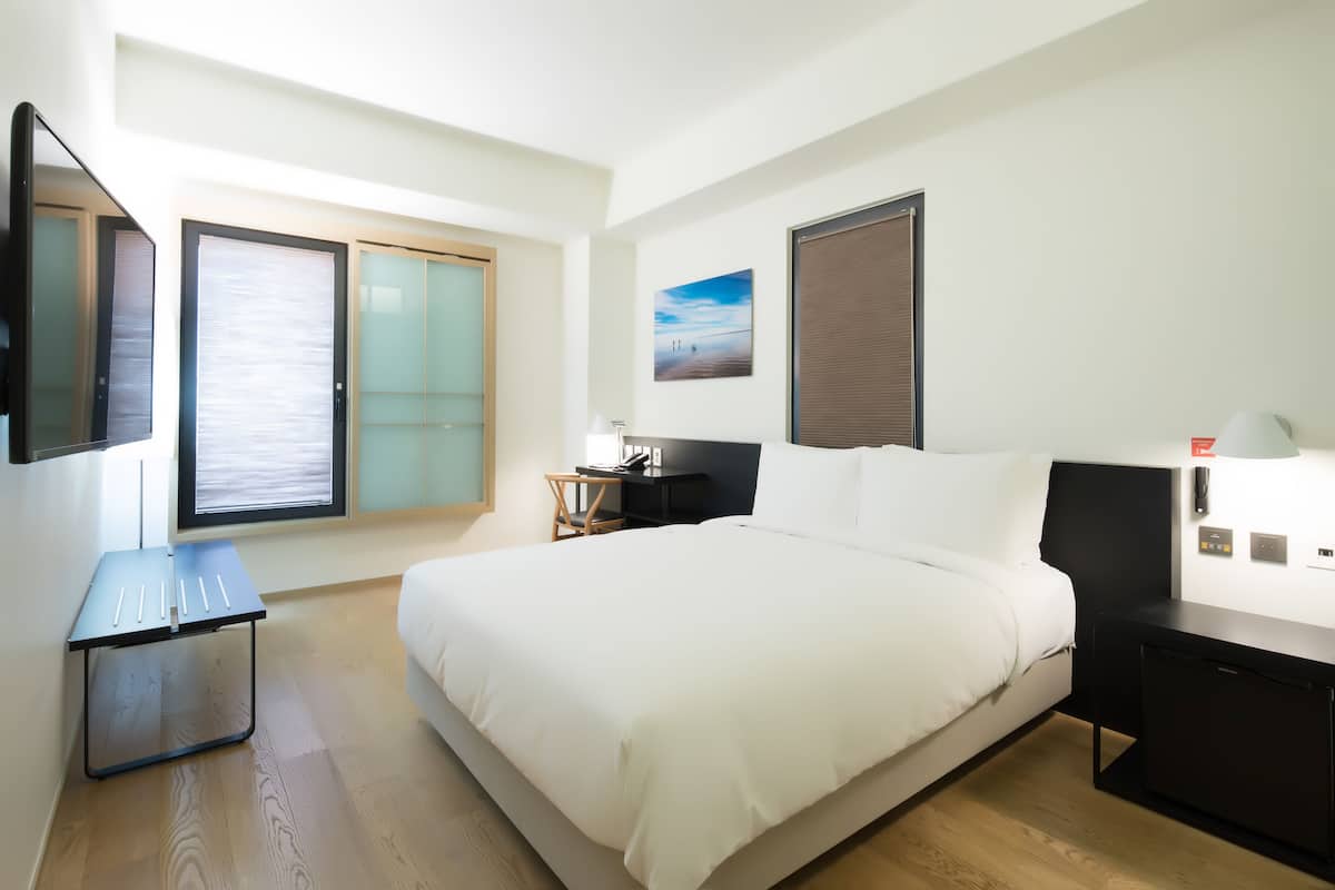 Ein modernes Hotelzimmer mit einem großen Bett, einem Schreibtisch mit Stuhl, einem kleinen Tisch, einem Fernseher und einem Fenster mit Jalousien.