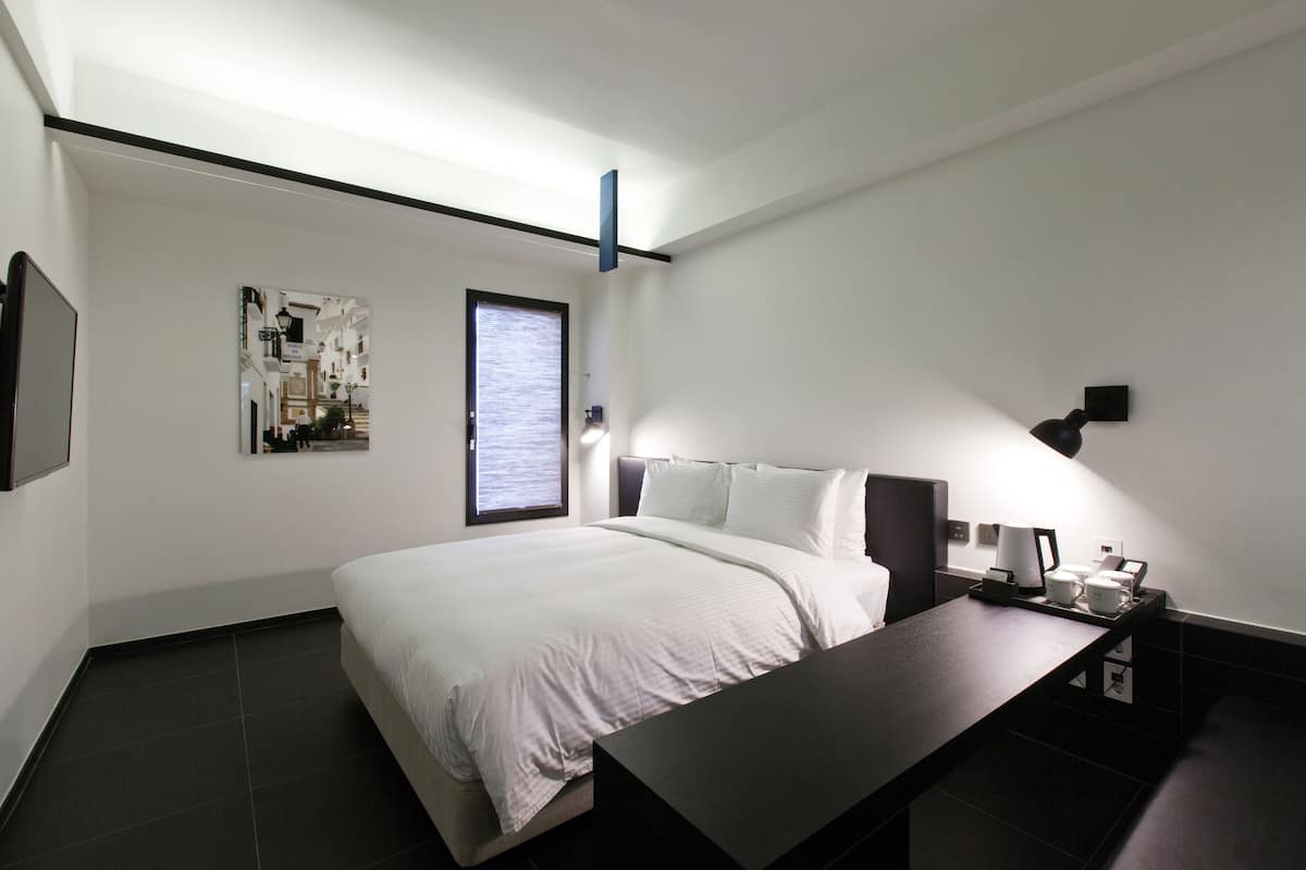 Ein modernes Hotelzimmer mit einem großen Bett, einem Nachttisch mit Lampe, einem kleinen Schreibtisch und einem Fenster mit Jalousien.