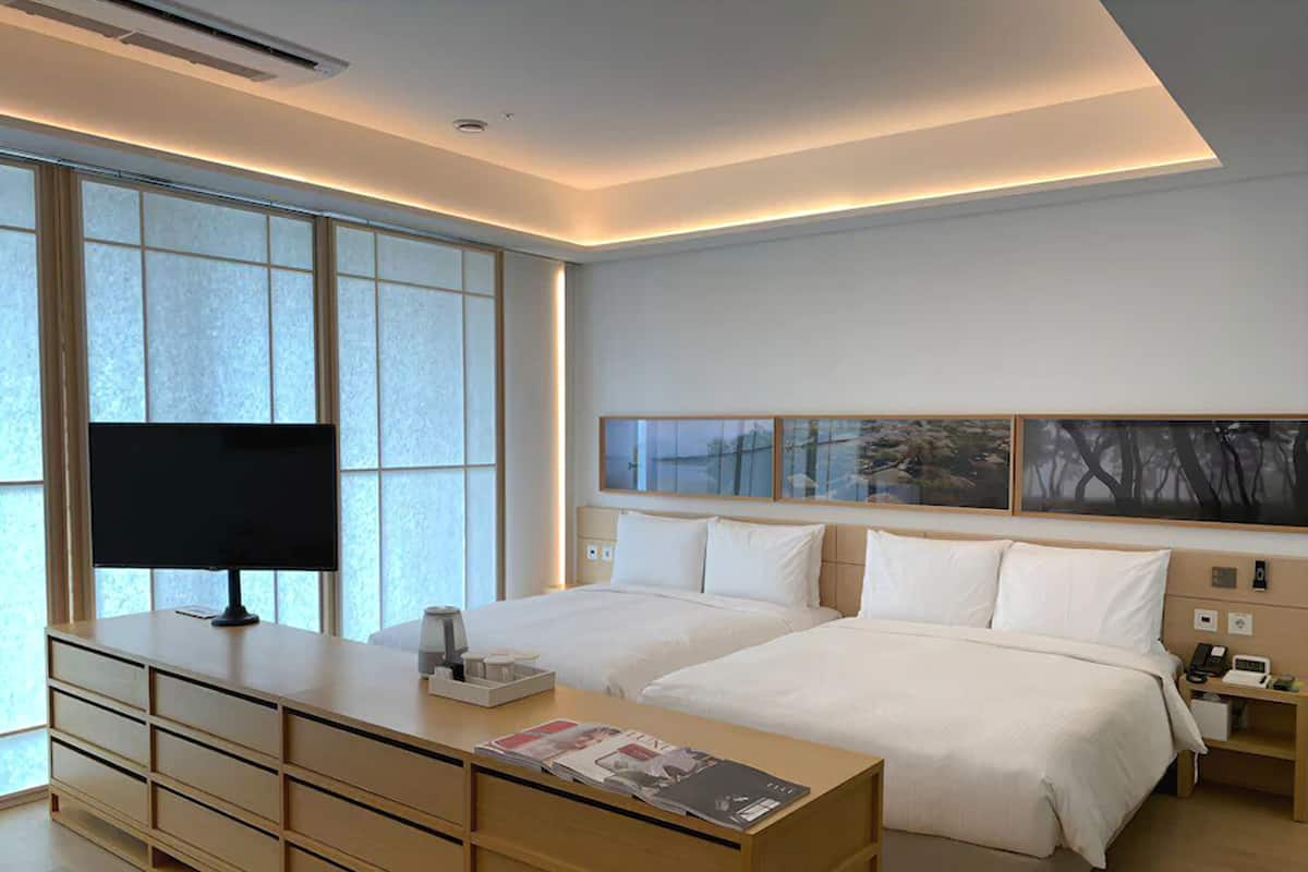 Ein modernes Hotelzimmer mit großem Fenster, einem Schreibtisch mit Computer, zwei Betten und Bildern an der Wand.