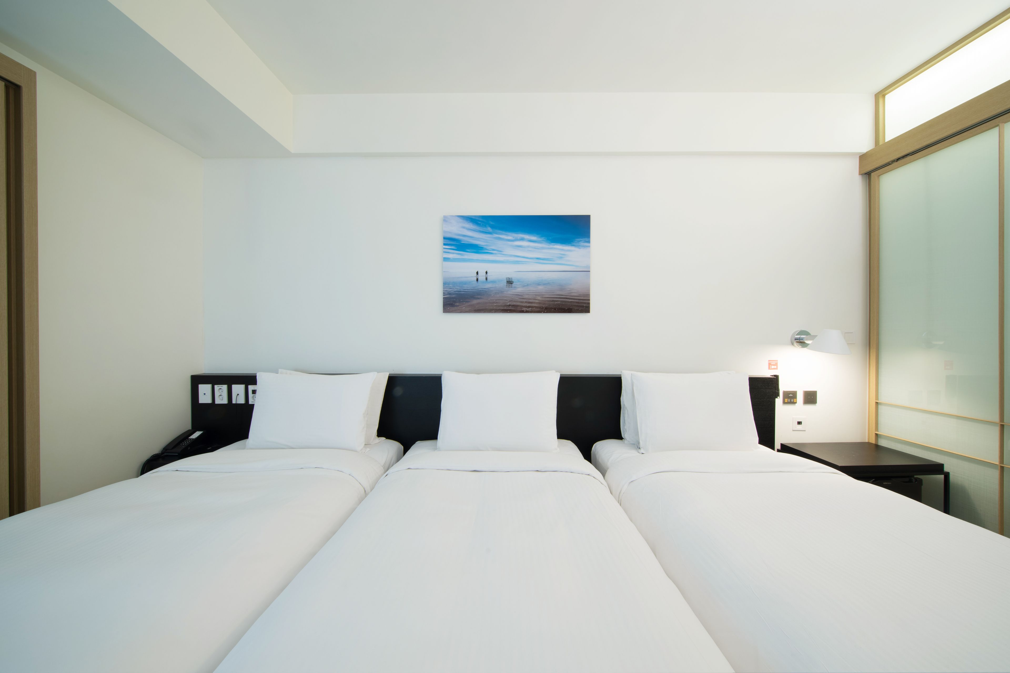 Una habitación de hotel con una cama grande, una mesita de noche, una lámpara y un cuadro de una playa en la pared.