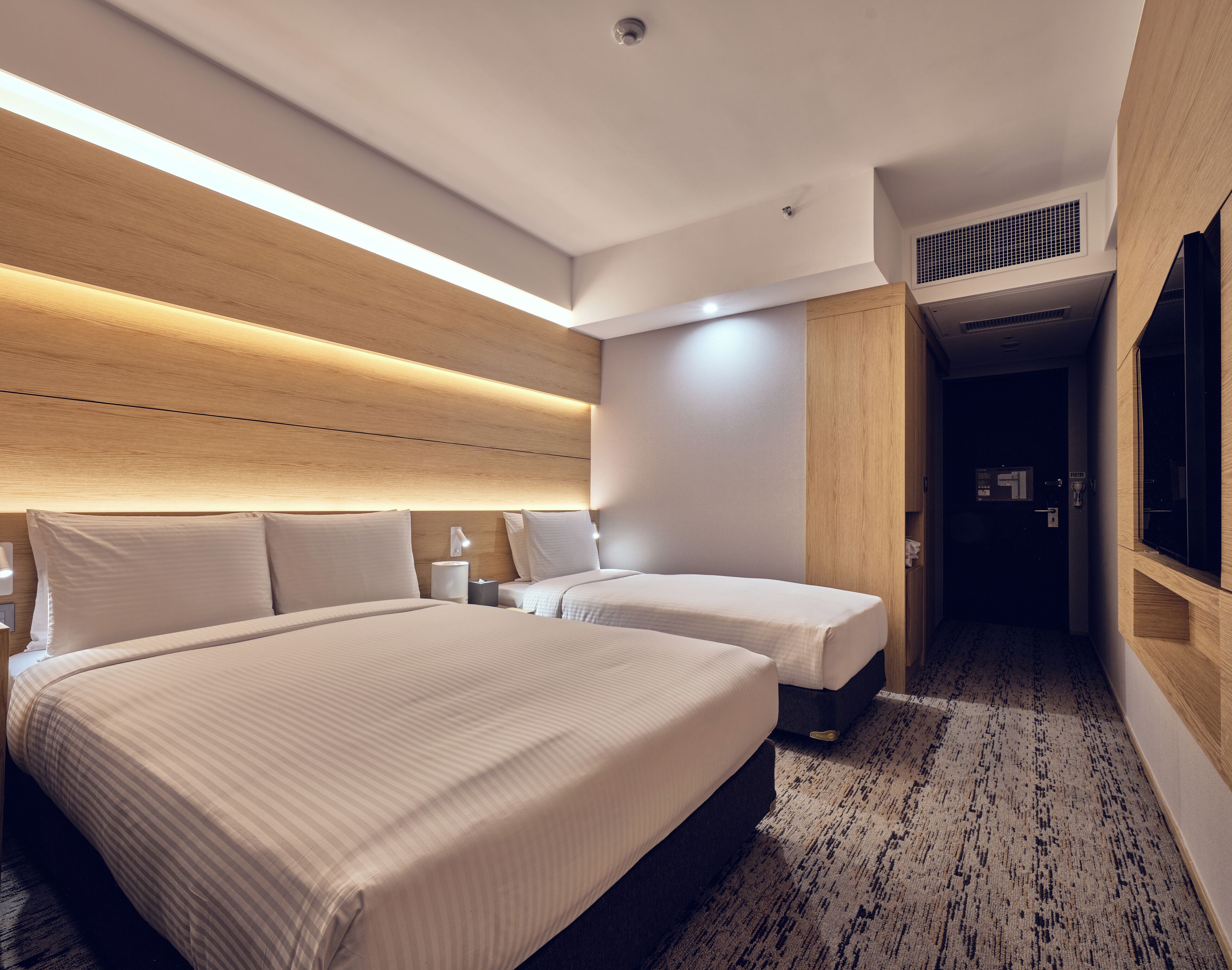 Habitación de hotel con dos camas, cabecero de madera, mesita de noche pequeña con lámpara y piso alfombrado.