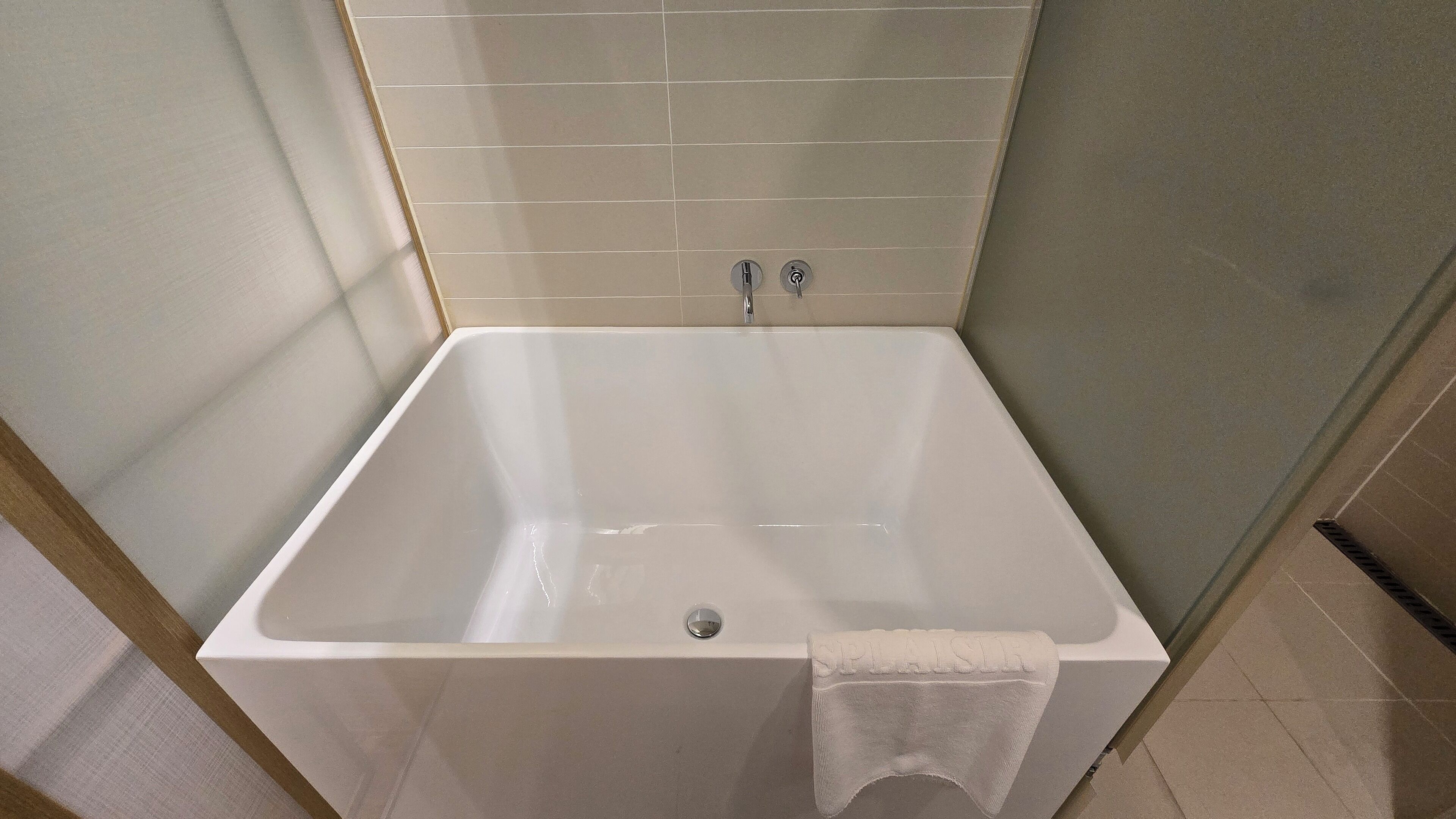 Uma banheira branca com uma toalha ao lado, parede revestida com azulejos e ralo.