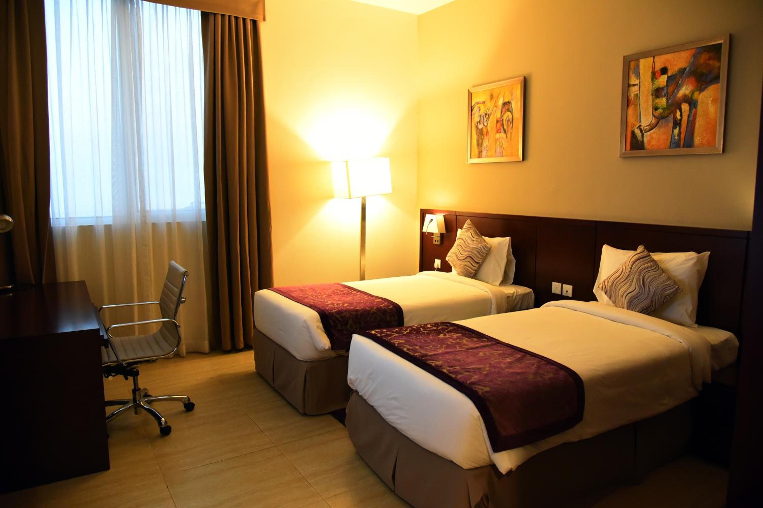 A hotel room with two beds, a desk, a chair, and a lamp.