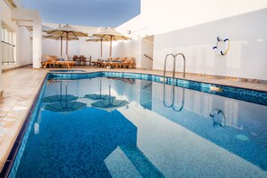 A swimming pool with lounge chairs and umbrellas, a tiled deck, and a lifebuoy on the wall. - V Hotel Fujairah (Al-Fujairah)