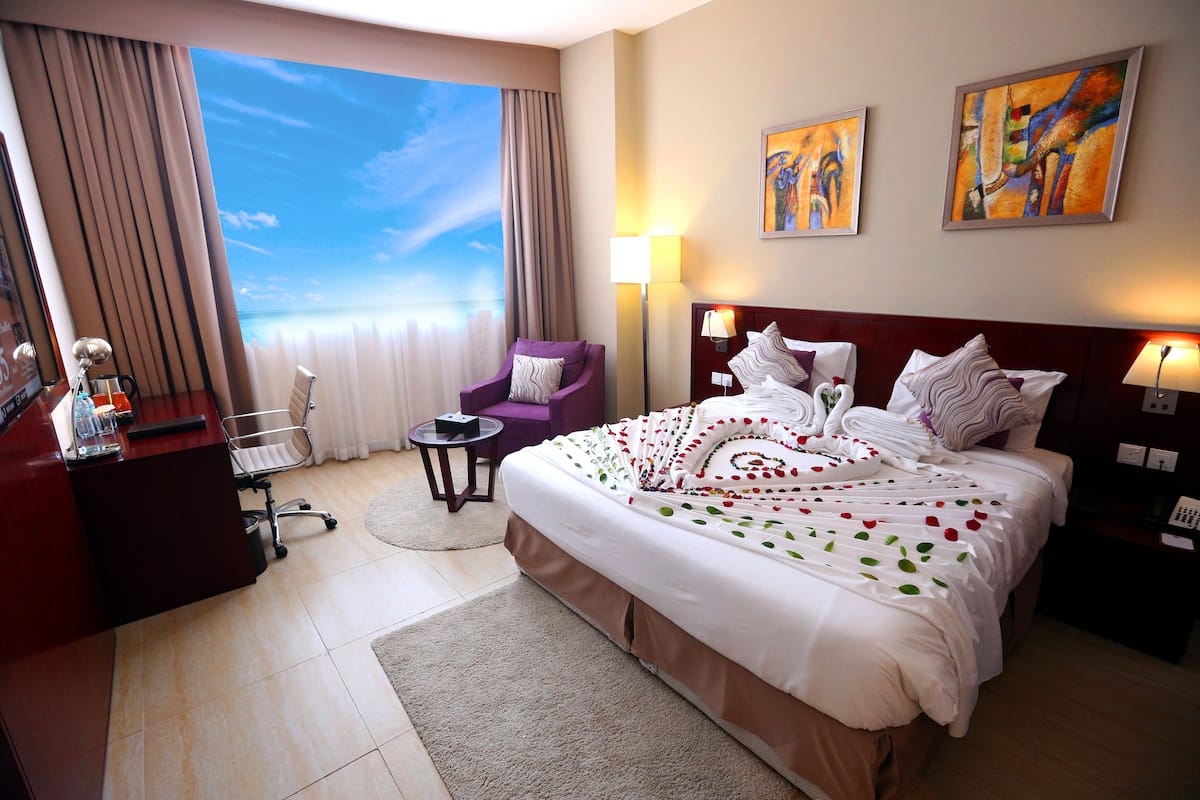 A hotel room with a large bed, a desk, a chair, and a view of the sky through the window.