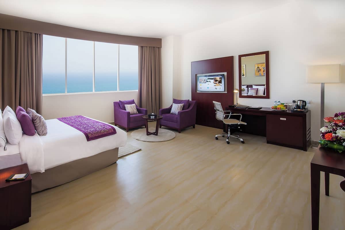 A hotel room with a large bed, two purple armchairs, a desk with a computer, and a view of the sea through large windows.