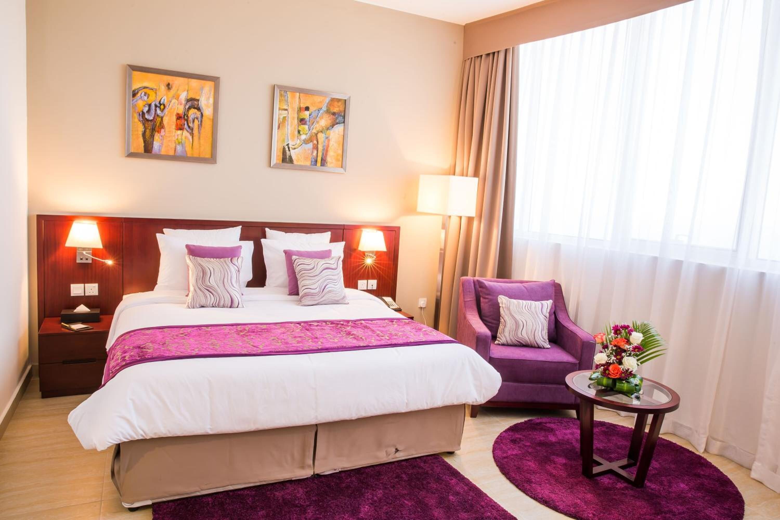 Quarto de hotel com uma cama grande, duas luminárias de cabeceira, uma poltrona roxa, uma mesa pequena com um vaso de flores e duas pinturas abstratas na parede.