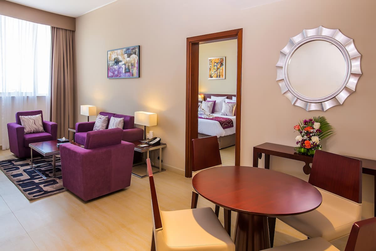 A hotel room with a purple sofa set, a wooden dining table, a mirror, and artwork on the walls.