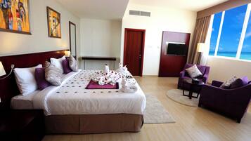 Habitación de hotel con una cama grande, zona de estar con una silla y una mesa, televisión y vista al océano.