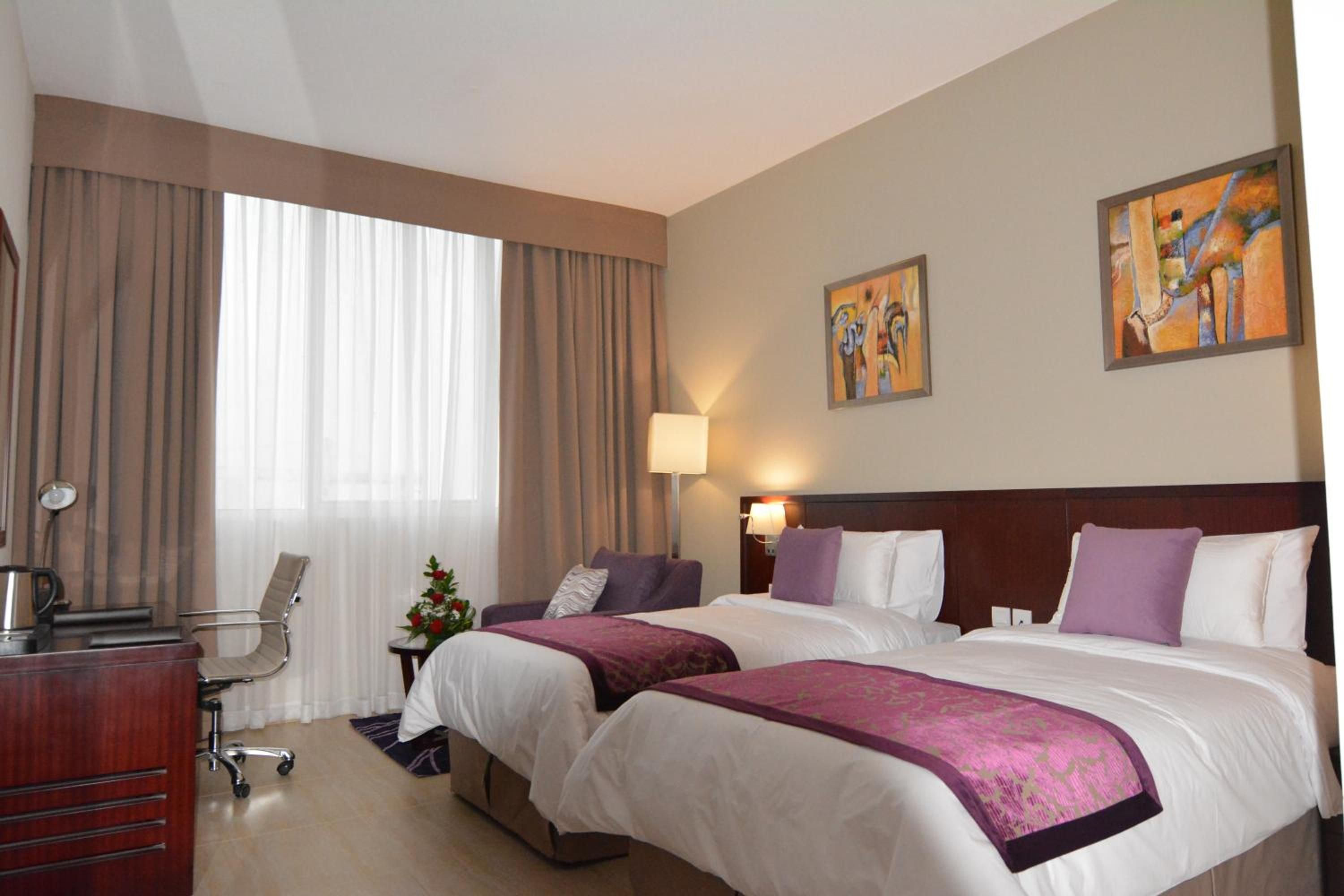 A hotel room with two beds, a desk, a chair, a lamp, and artwork on the walls.