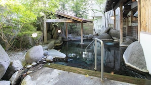Un onsen japonés tradicional con estructura de madera, asientos de piedra y una piscina de agua cristalina rodeada de rocas y vegetación.