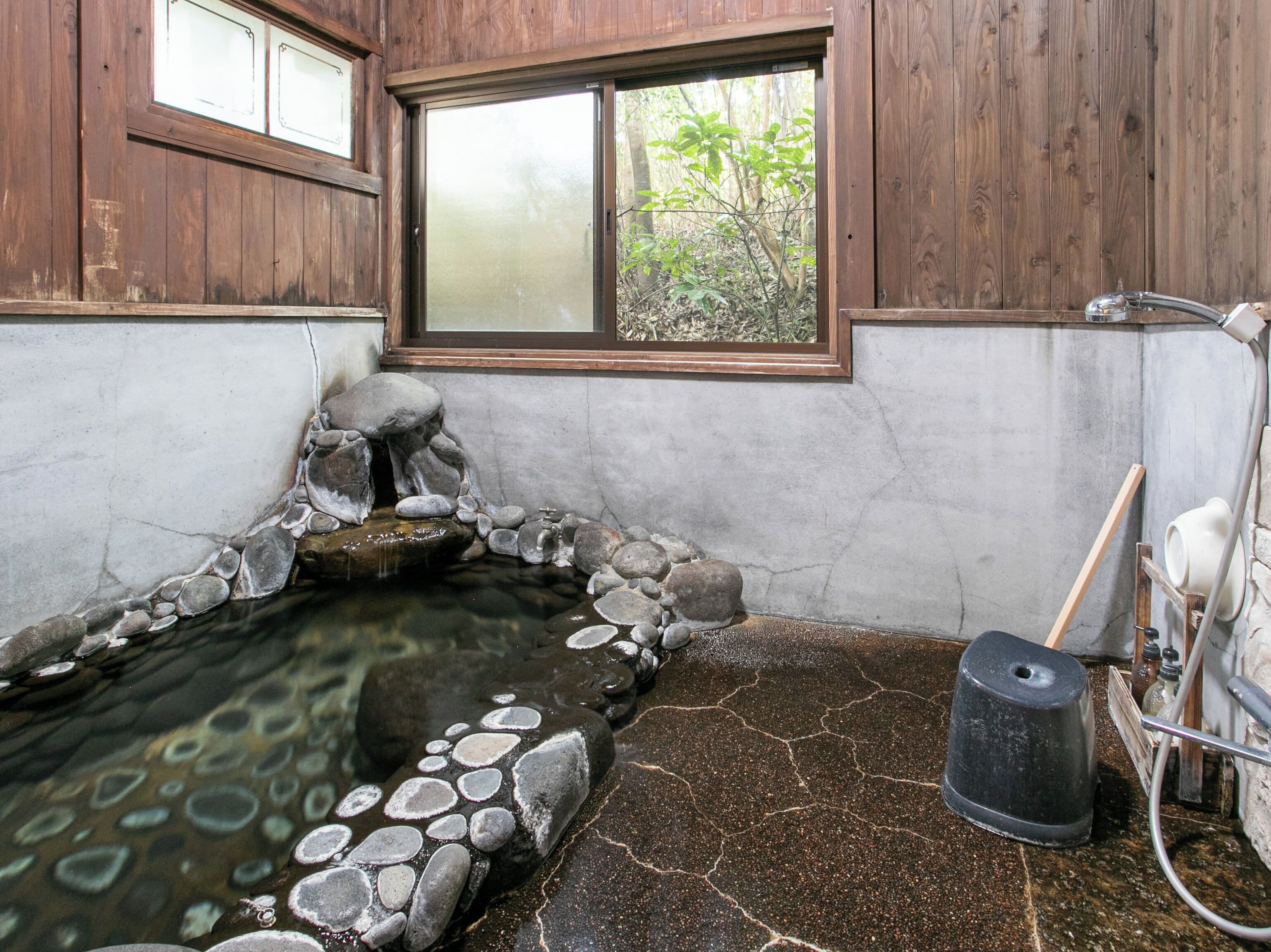 Um onsen japonês tradicional com banheira de pedra, paredes de madeira e vista para o verde lá fora.