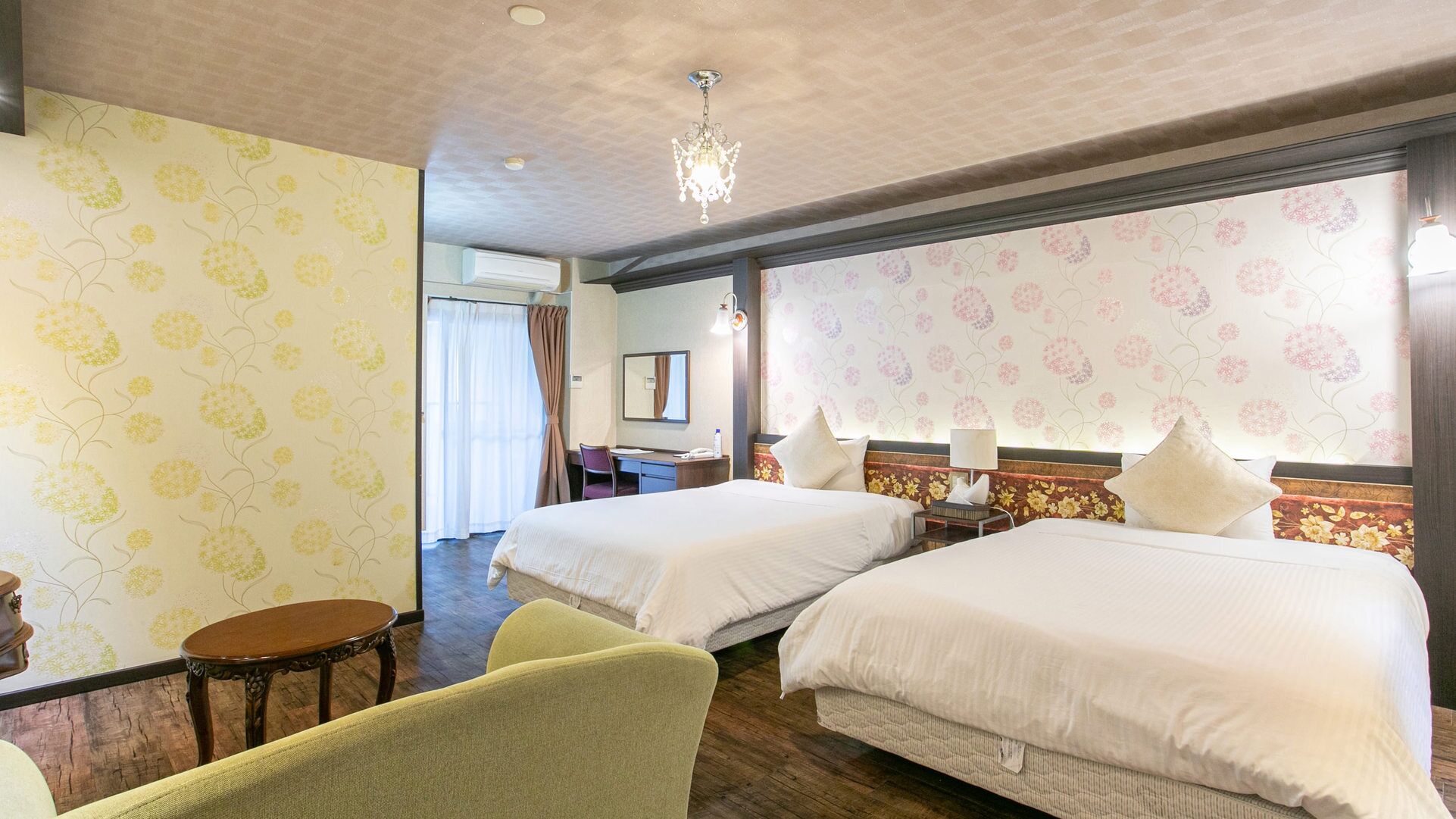 Une chambre d’hôtel avec deux lits, un bureau, une chaise, un lustre et un papier peint à motifs floraux.