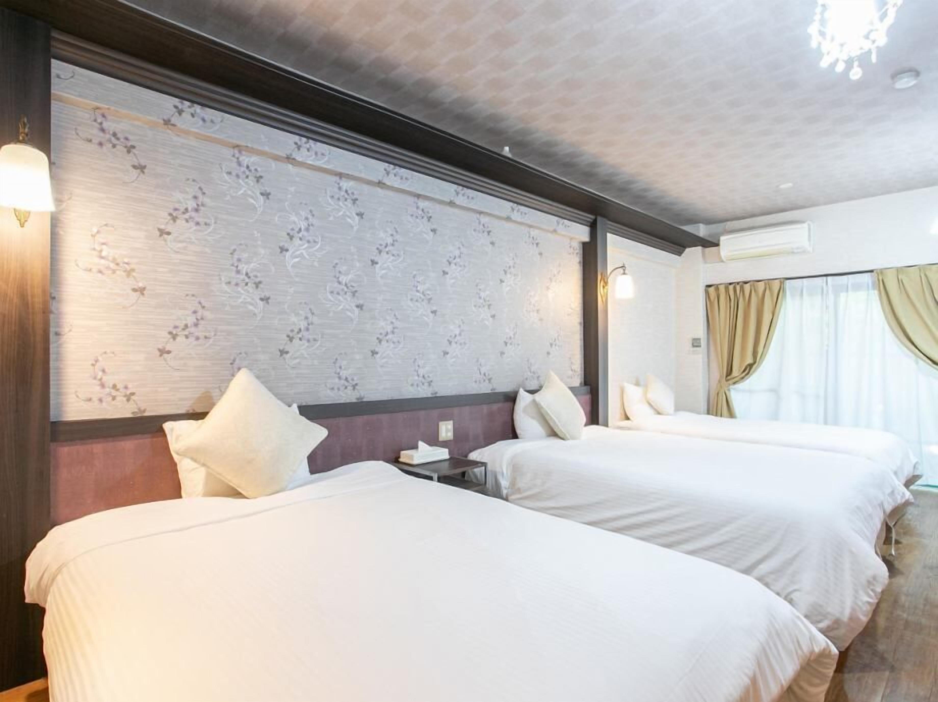 A hotel room with two beds, a large window with curtains, a wall with a floral pattern, and a chandelier.