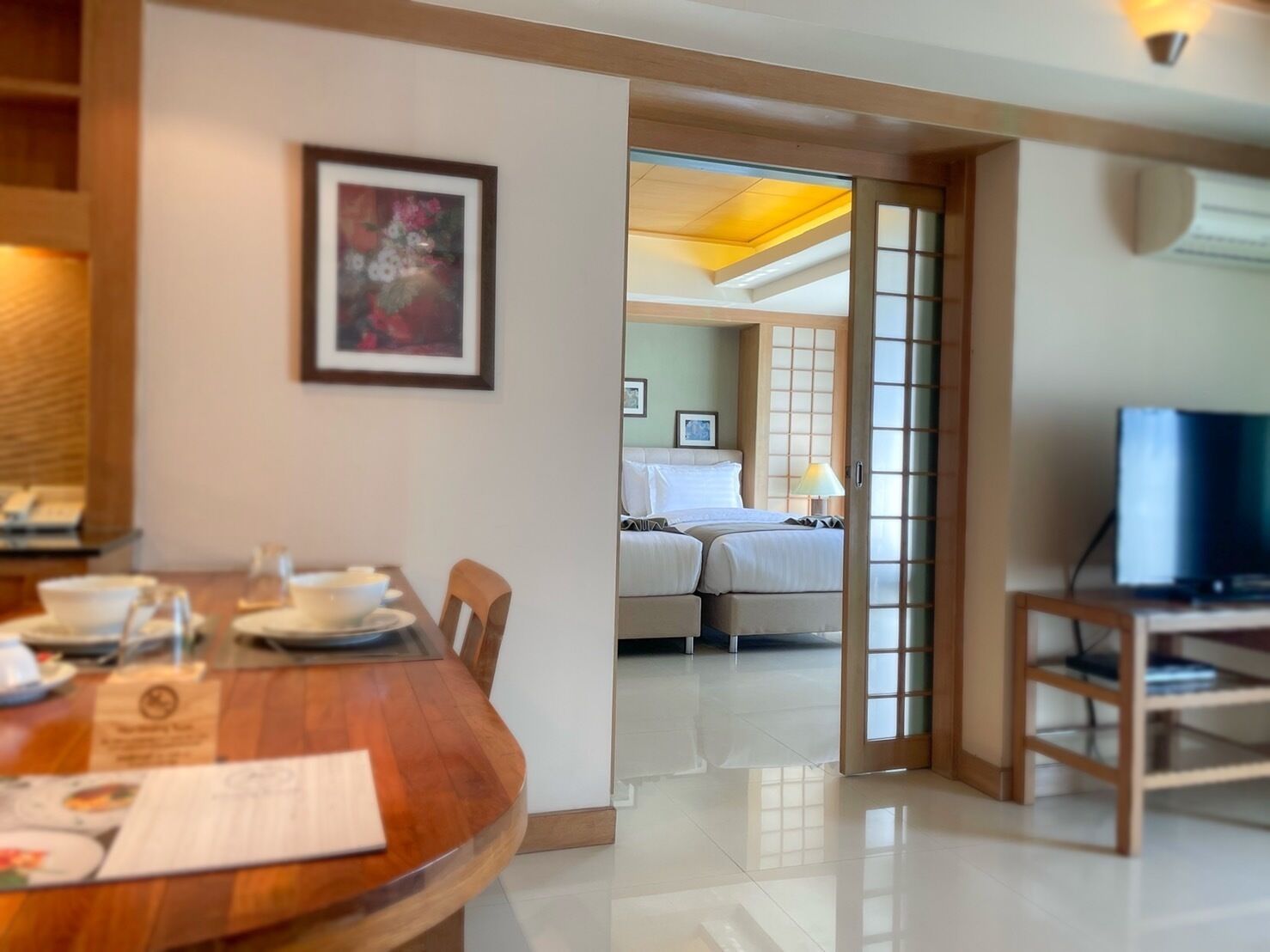 A hotel room with a wooden dining table, a bed with white linens, a television on a stand, and a sliding door leading to another room.