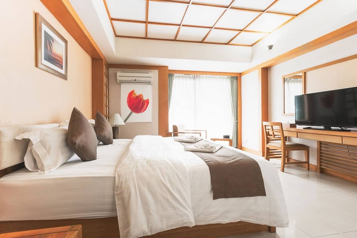 A hotel room with a bed, a desk with a TV, a window with curtains, and a wall-mounted air conditioner.