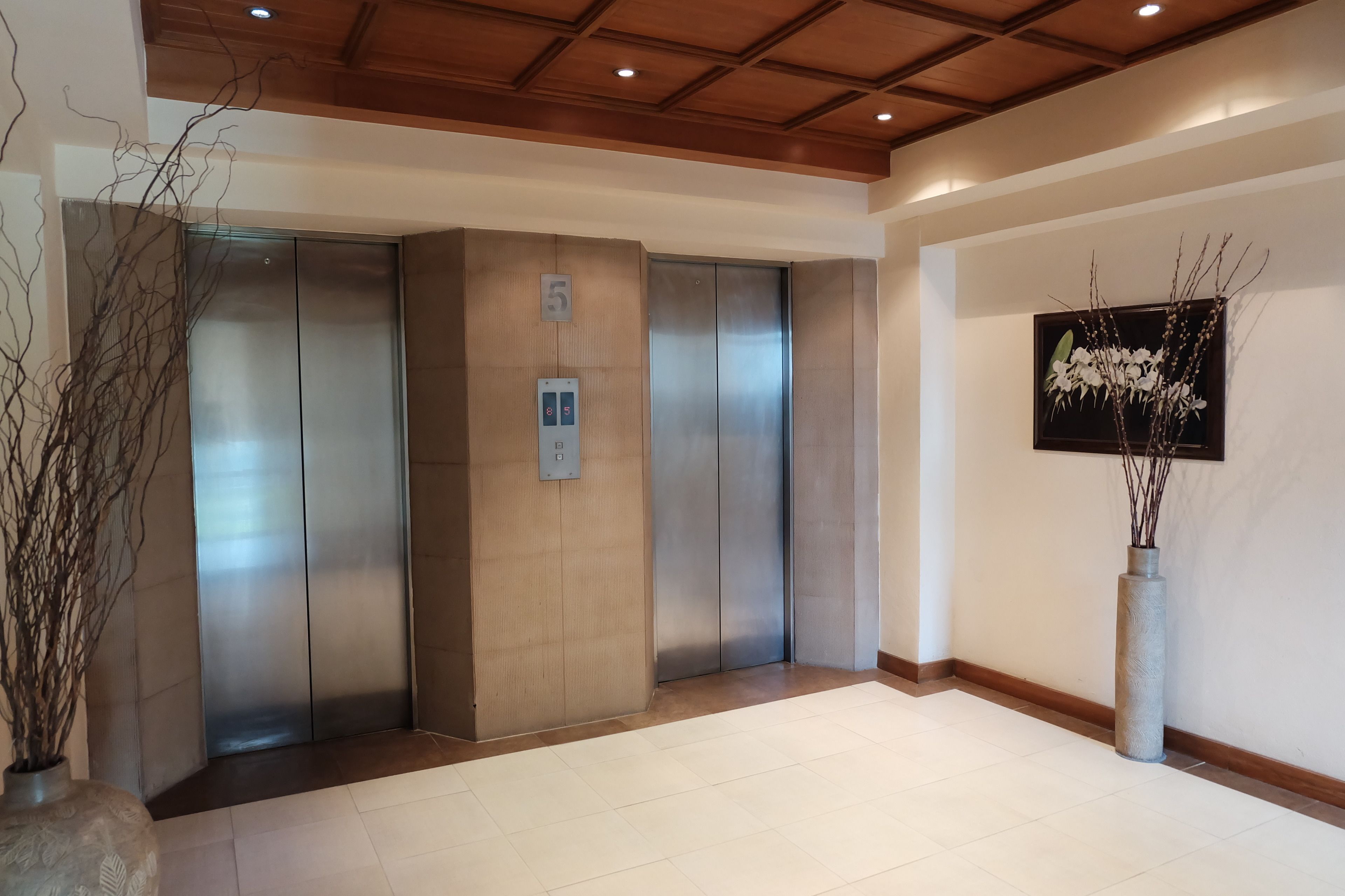 A modern elevator area with a wooden ceiling, a wall-mounted elevator control panel, and a vase with branches.