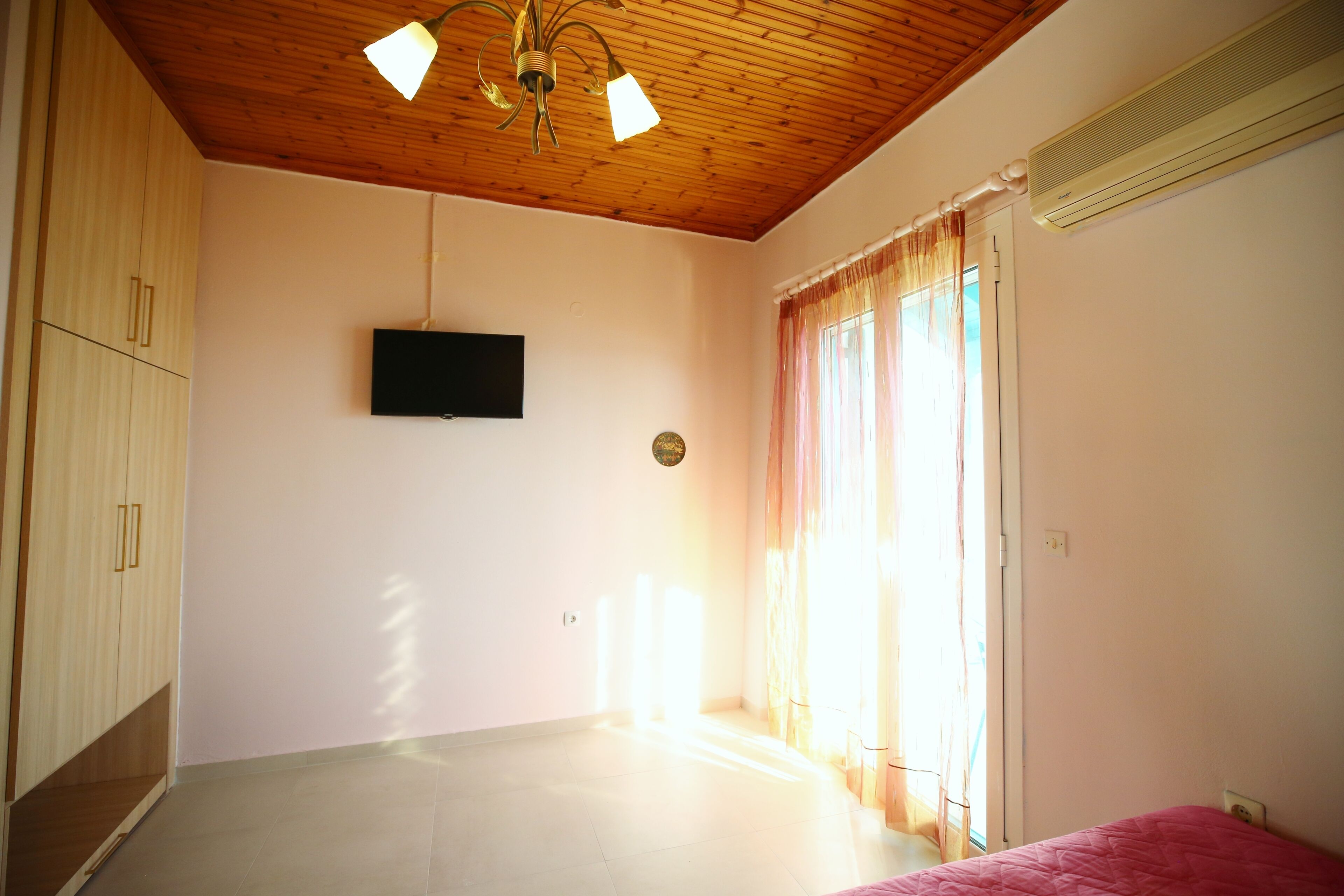 A room with a wooden ceiling, a flat-screen TV mounted on the wall, and a bed with a pink cover.