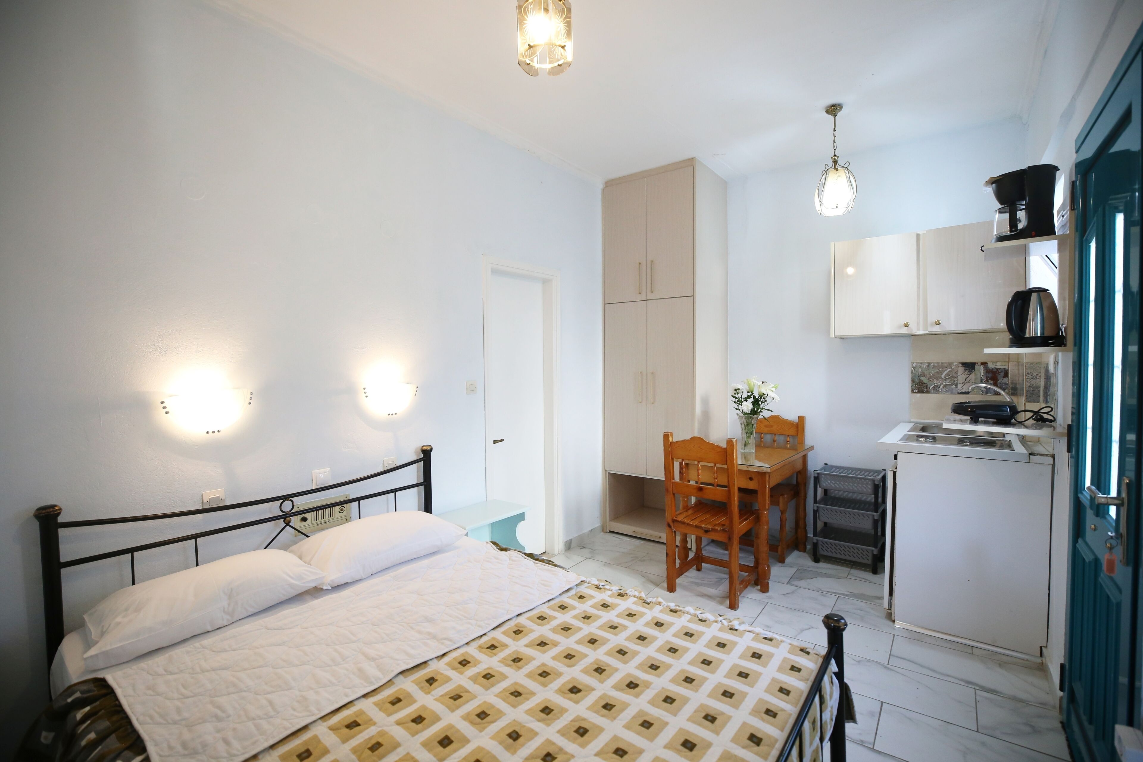 A compact room with a bed, a small table, a chair, a kitchenette, and a door.