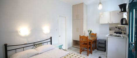 A compact room with a bed, a small table, a chair, a kitchenette, and a door.