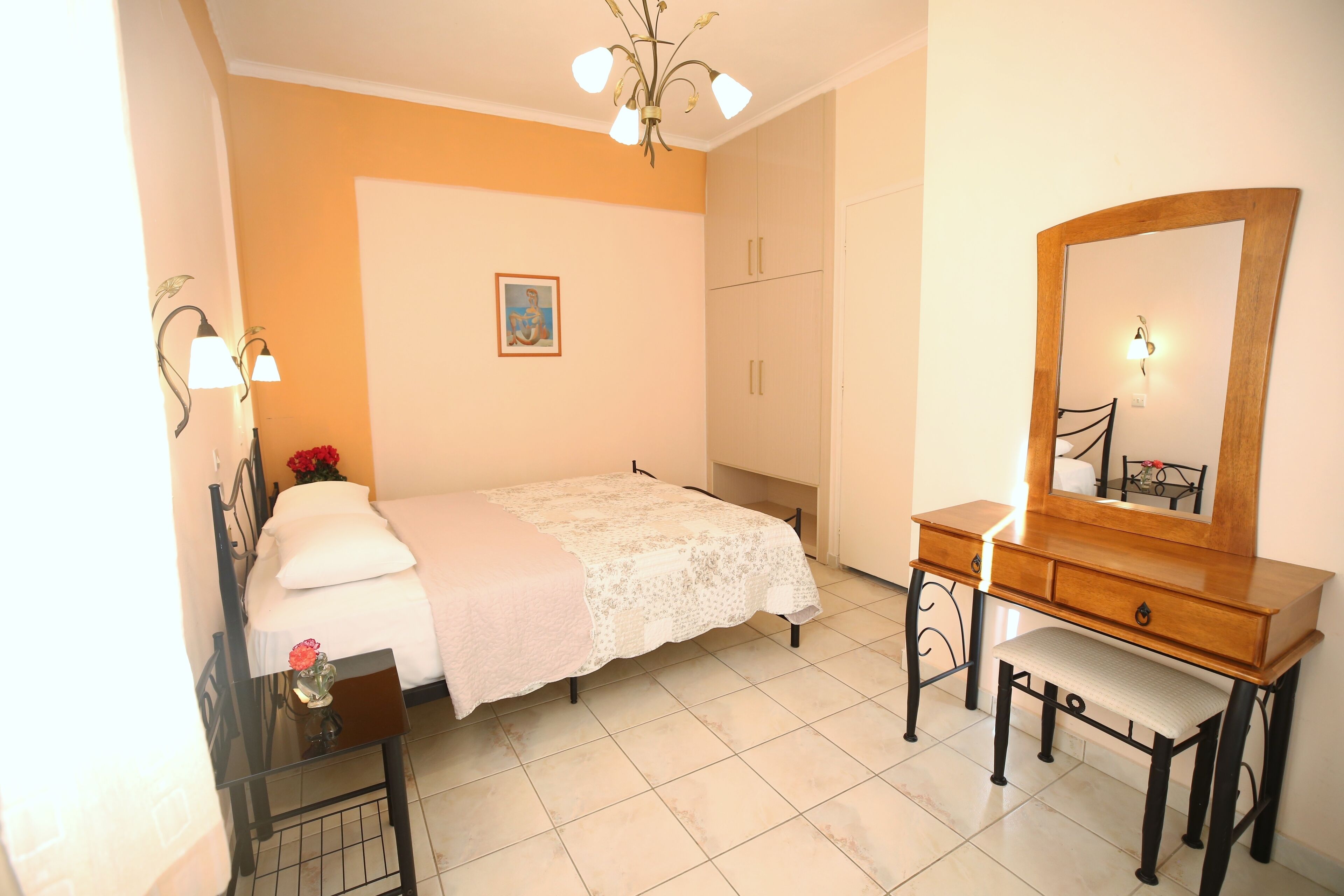 A bedroom with a bed, a wooden dresser, a bench, a nightstand, a mirror, and a wall-mounted light fixture.