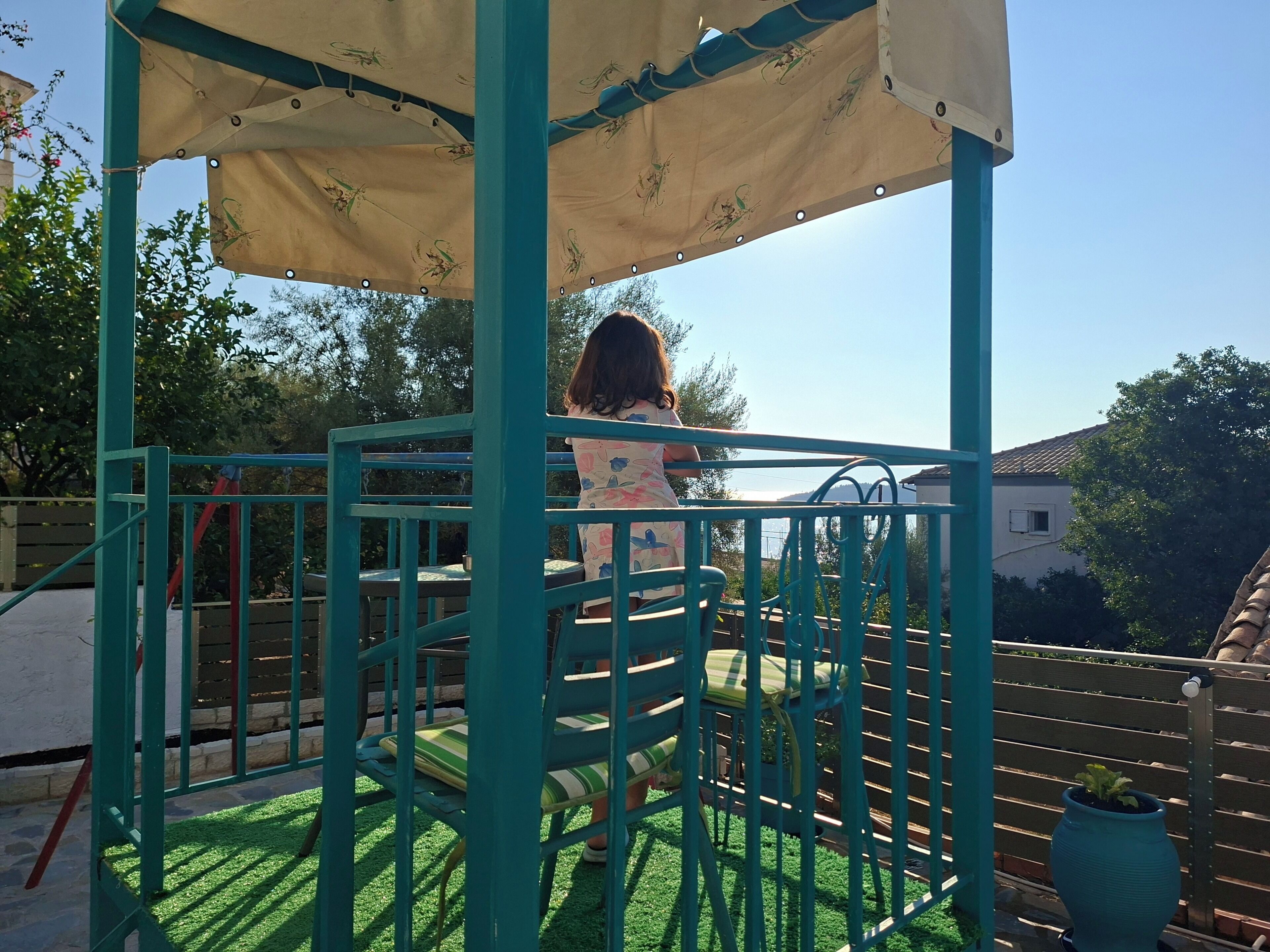 Niño en una estructura de juego verde con un dosel color crema, rodeado de una cerca y follaje.
