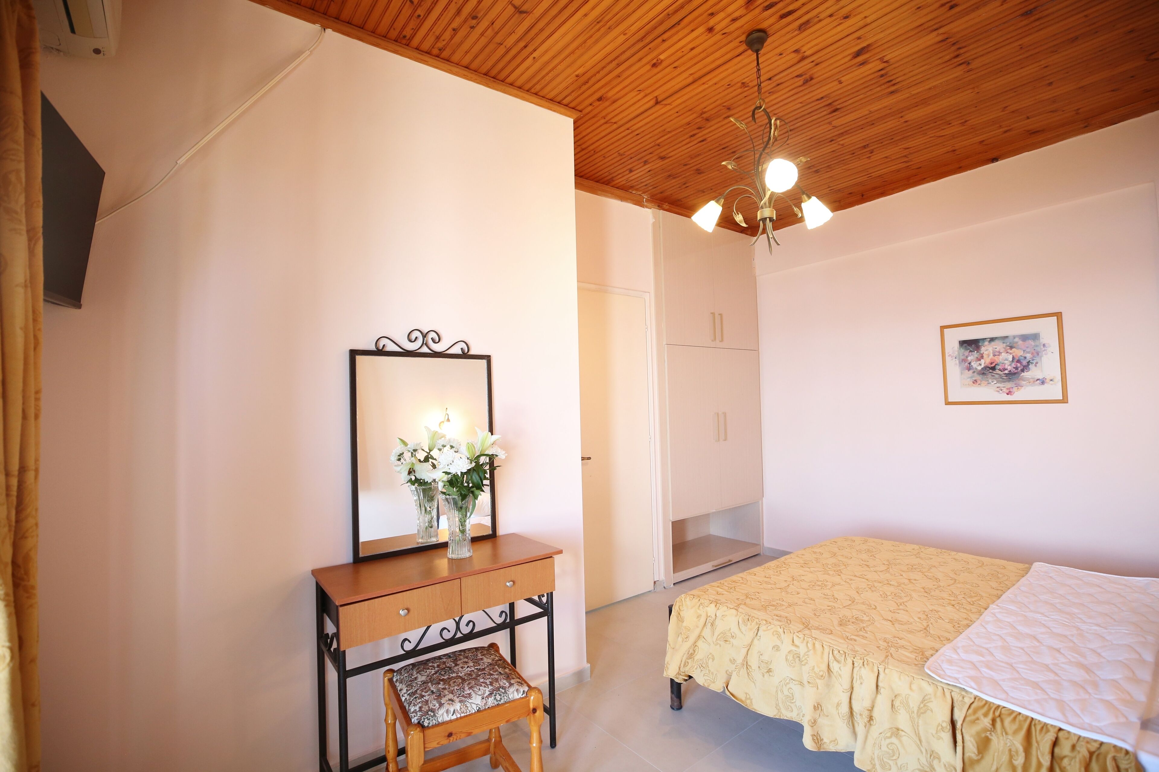 A bedroom with a wooden ceiling, a bed with a yellow bedspread, a nightstand with a mirror and vase, and a door leading to another room.