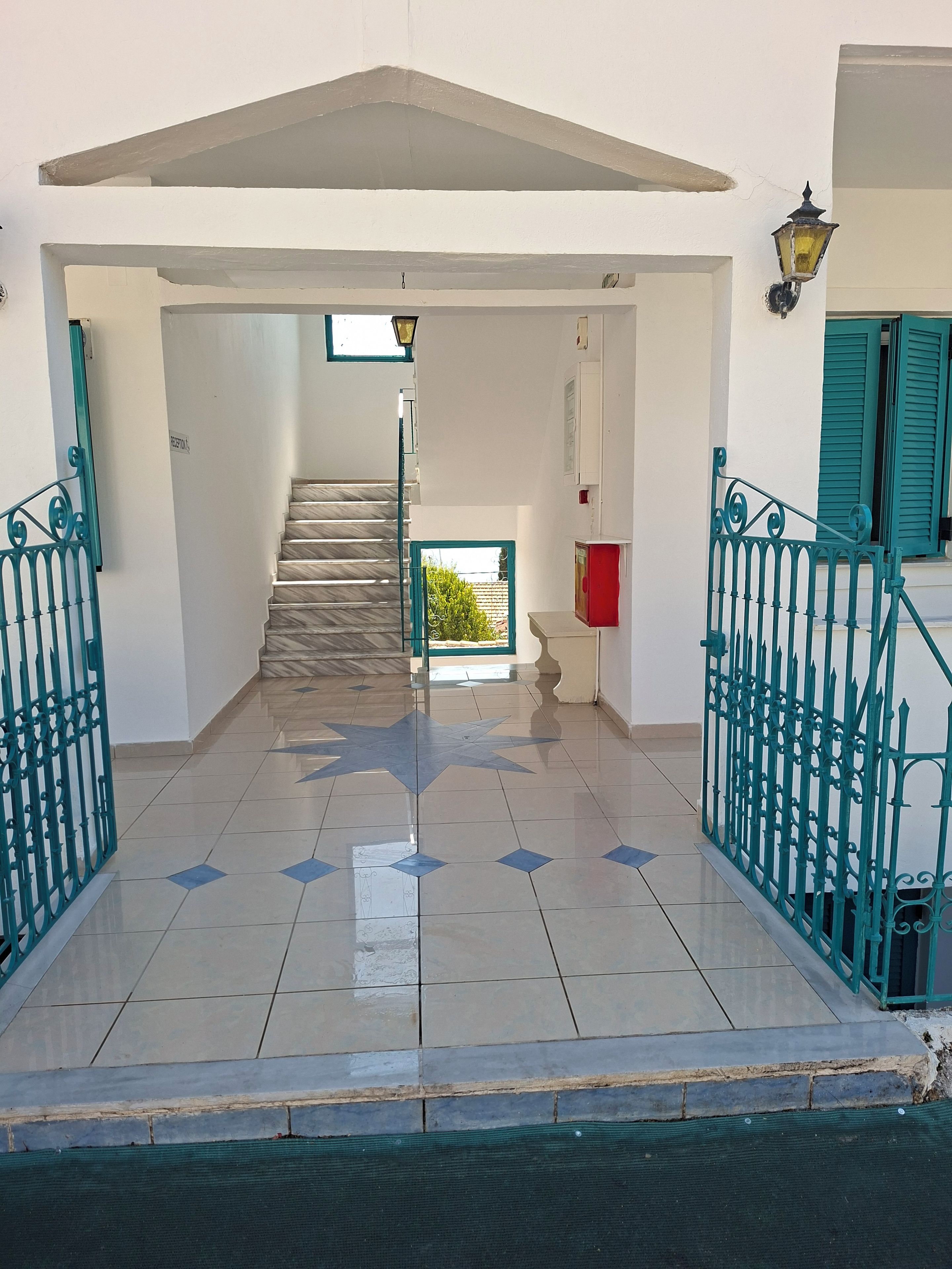 Un escalier menant à l’étage supérieur, un hall d’entrée carrelé et un portail en métal vert.