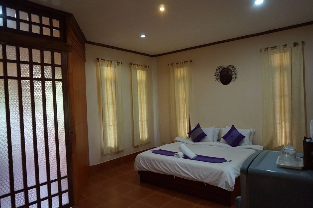 A hotel room with a bed, curtains, a wall-mounted decoration, and a small table with a glass object.