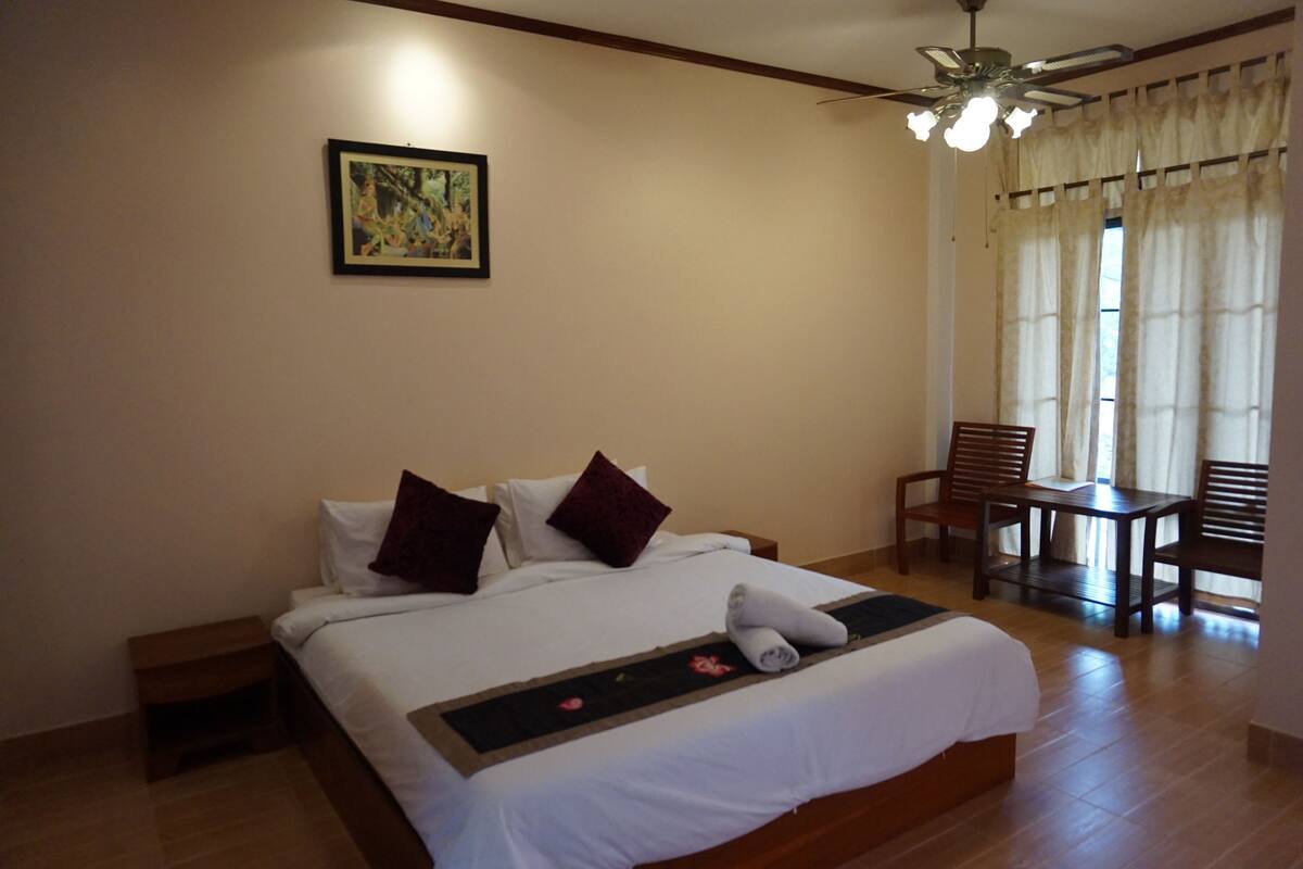 A neatly made bed with white linens and two pillows, a small bedside table, a ceiling fan, and a window with curtains.
