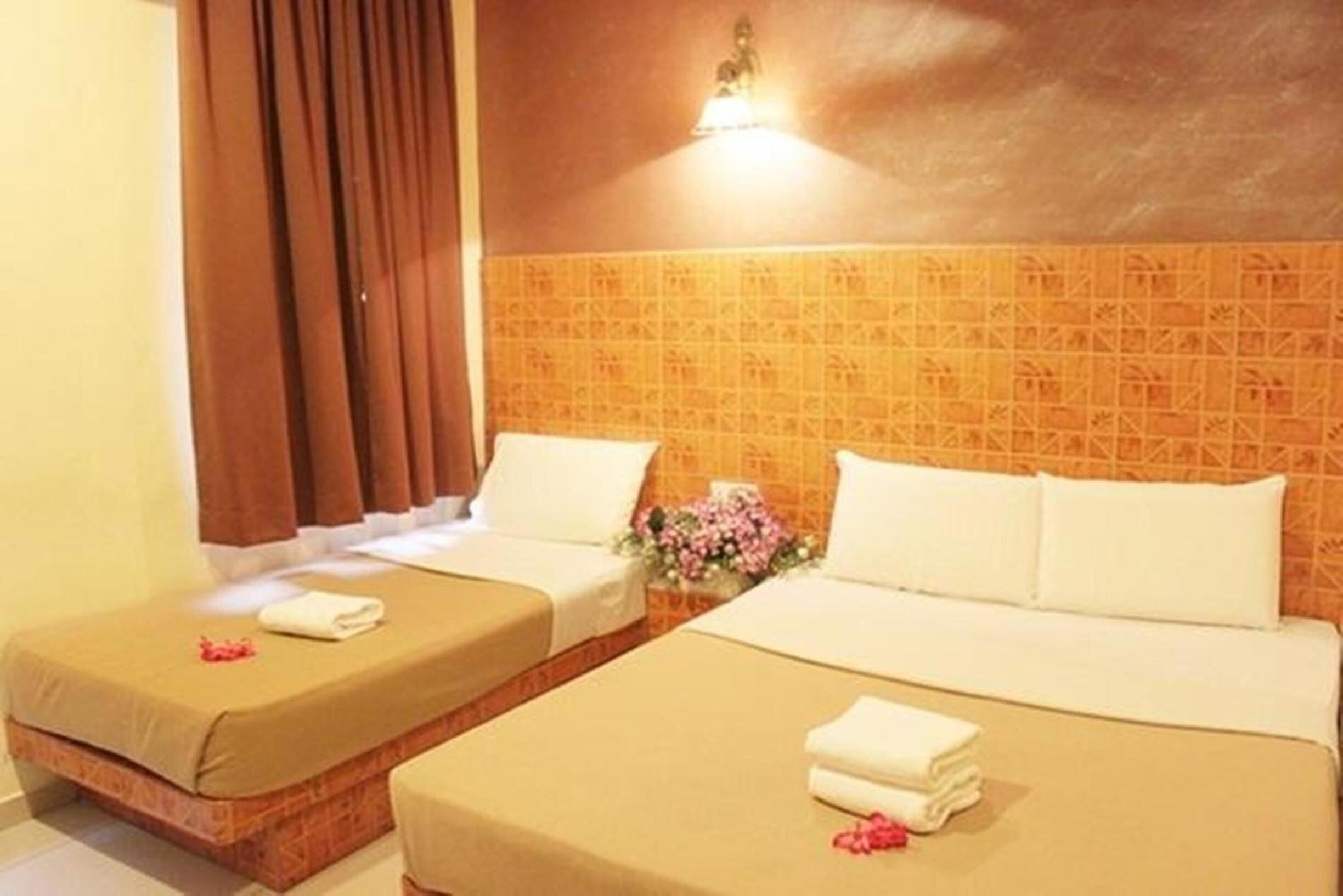 Habitación de hotel con dos camas, cada una con cubrecama color beige y almohadas blancas. Entre las camas hay una pequeña mesa con un florero de flores rosadas.