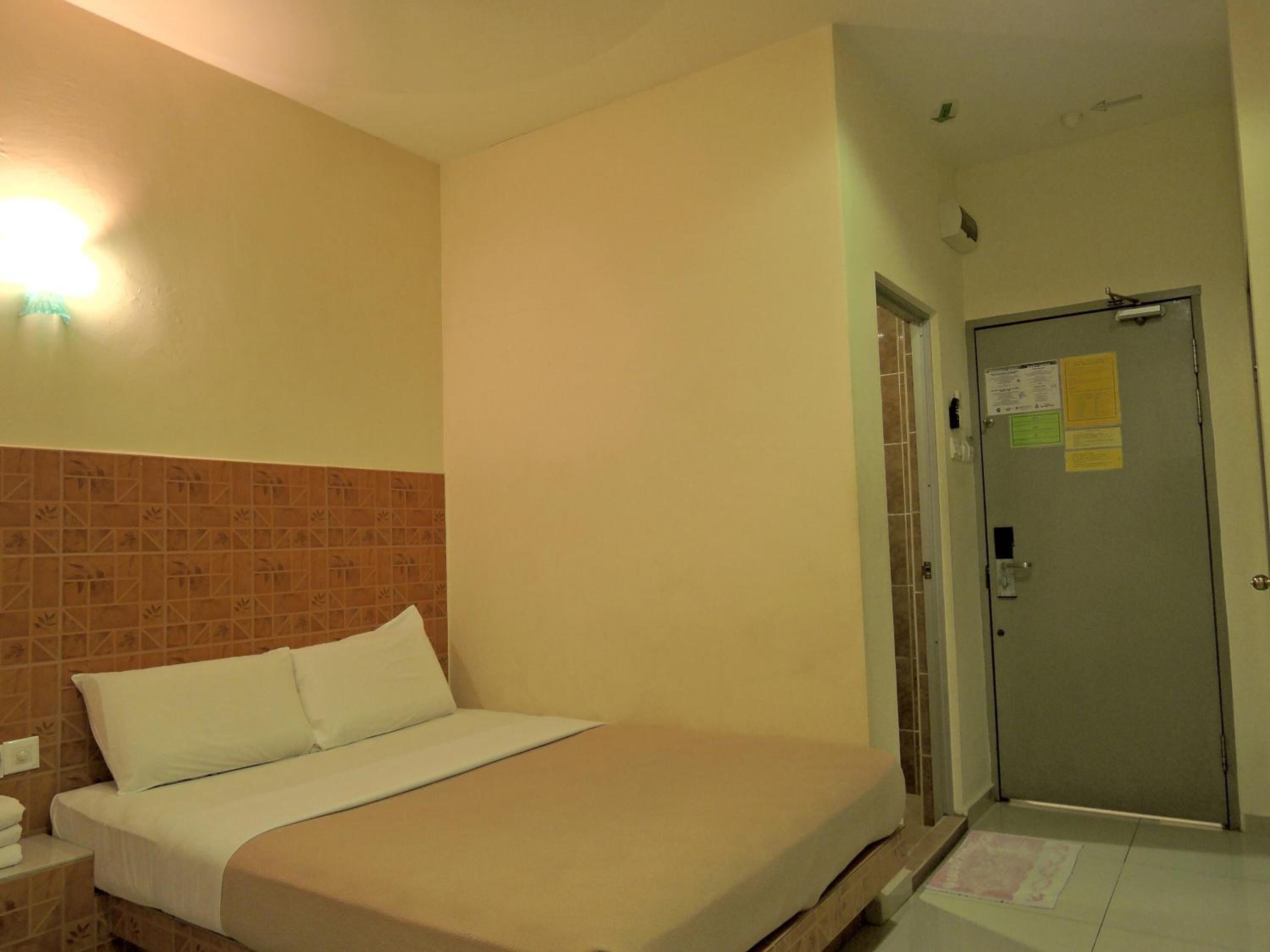 Habitació d'hotel amb un llit, una tauleta de nit, un capçal de llit amb rajoles, una porta amb un tancament i una lluminària a la paret.