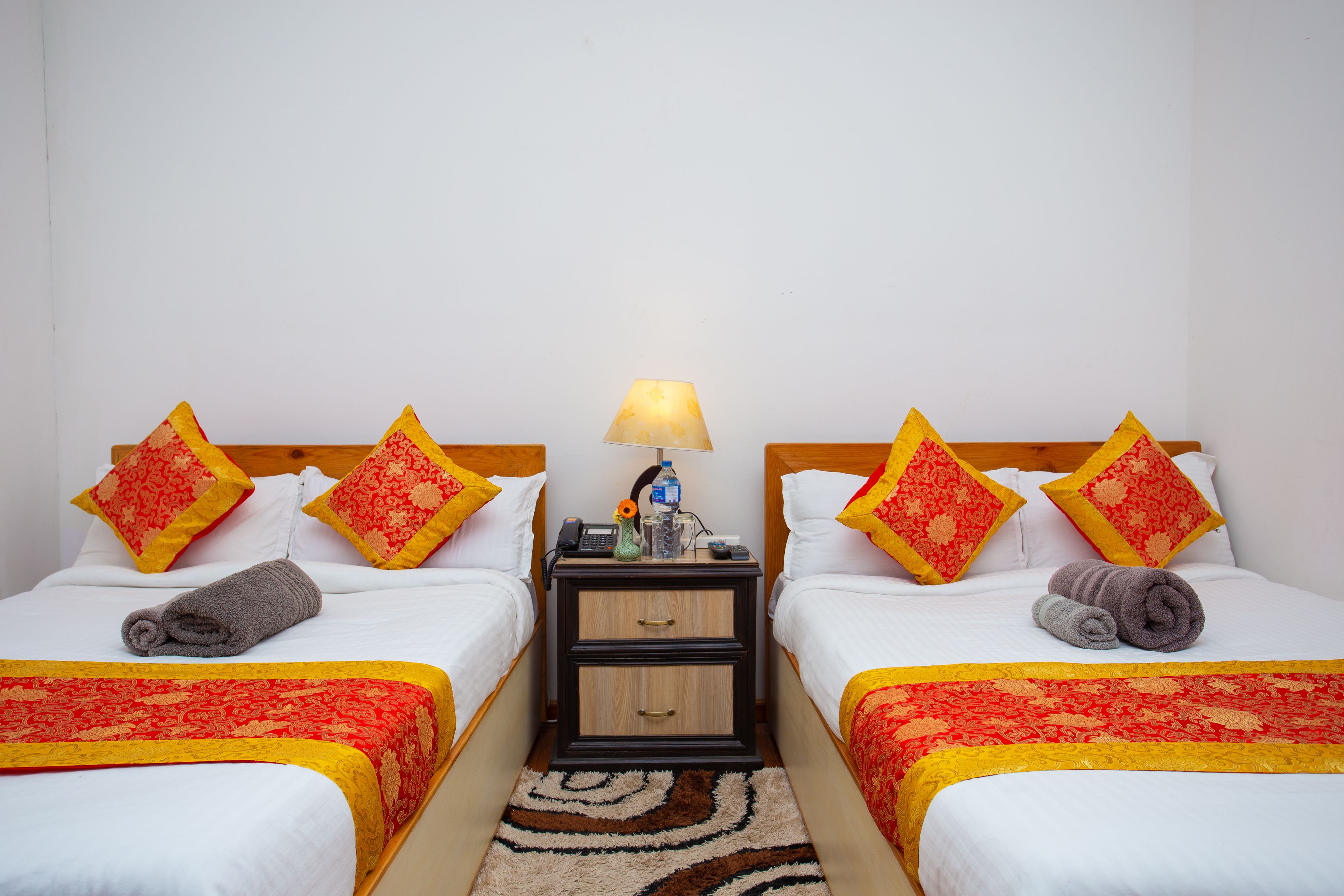 Een hotelkamer met twee bedden, elk voorzien van een rood-geel geboeide spreidek en witte kussens, evenals bijpassende sierkussens. Tussen de bedden staat een nachtkastje met een lamp, een telefoon en een waterfles.