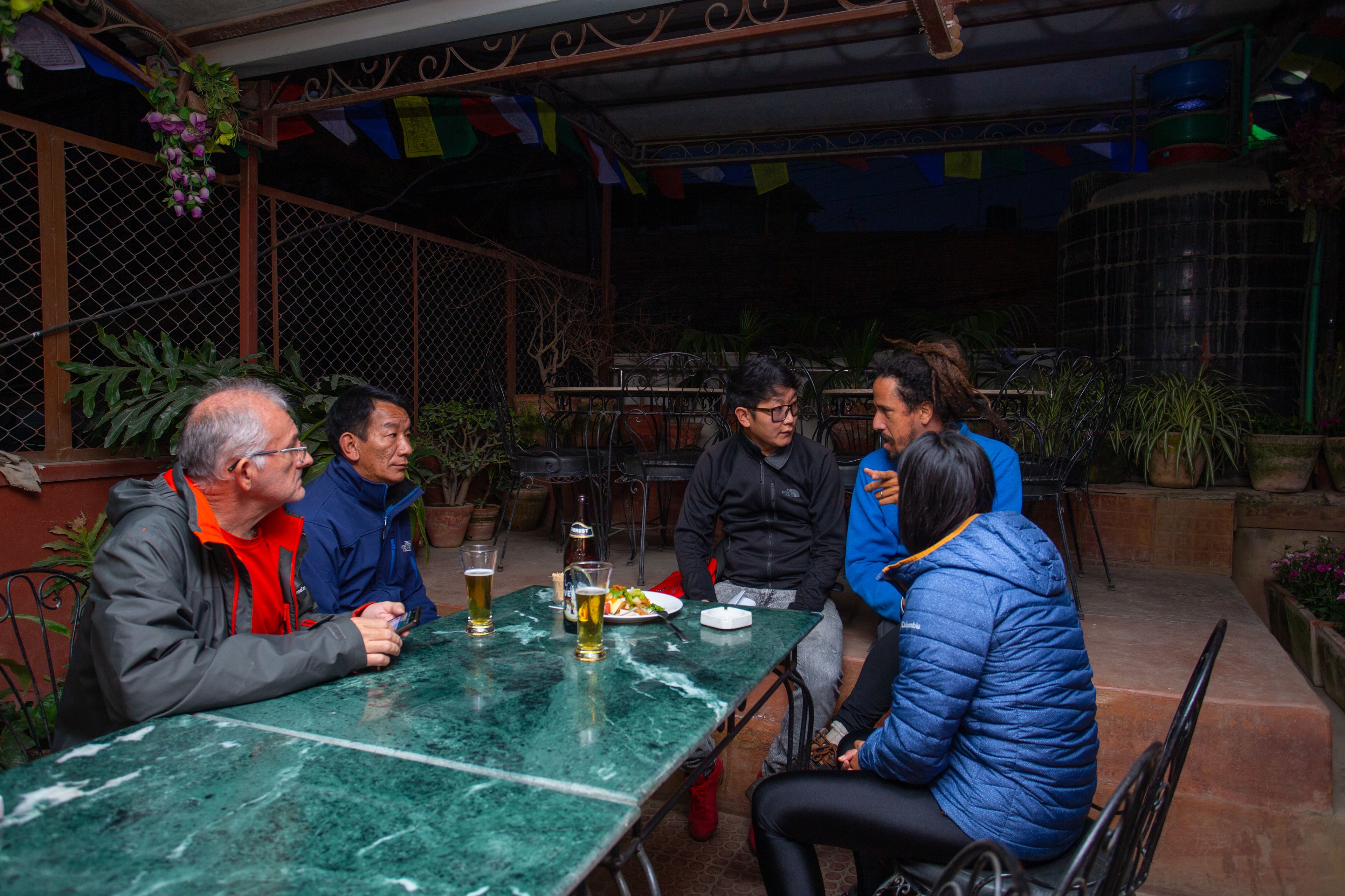 A group of people seated around a table with drinks, in an outdoor setting with plants and a metal fence.