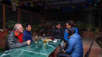 A group of people seated around a table with drinks, in an outdoor setting with plants and a metal fence.