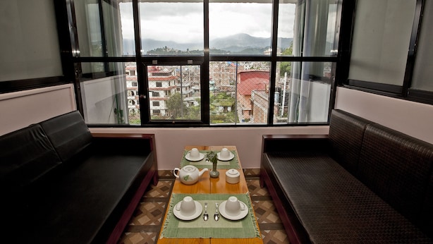 A dining area with a table set for two, a view of a cityscape through large windows, and a seating area with a bench. - The Boudha Inn (Kathmandu)