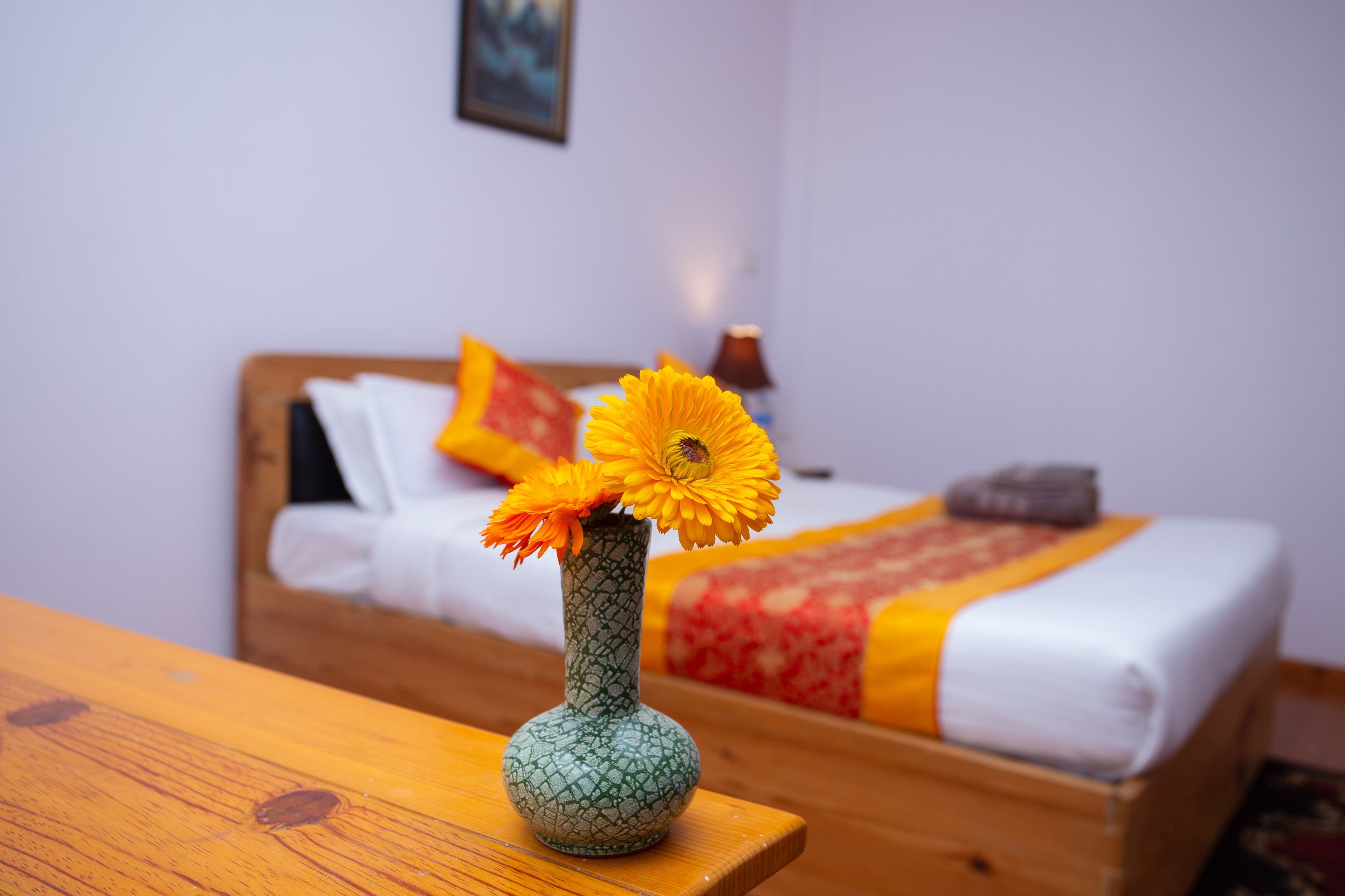 Una mesa de madera con un jarrón verde con flores amarillas, una cama con una colcha roja y amarilla, y una almohada blanca al fondo.