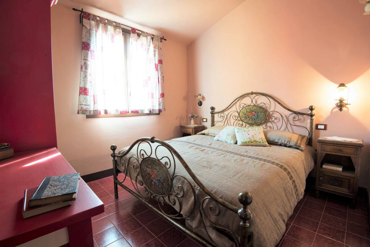 Un dormitorio con estructura de cama de metal, mesita de noche, lámpara de noche y ventana con cortinas.