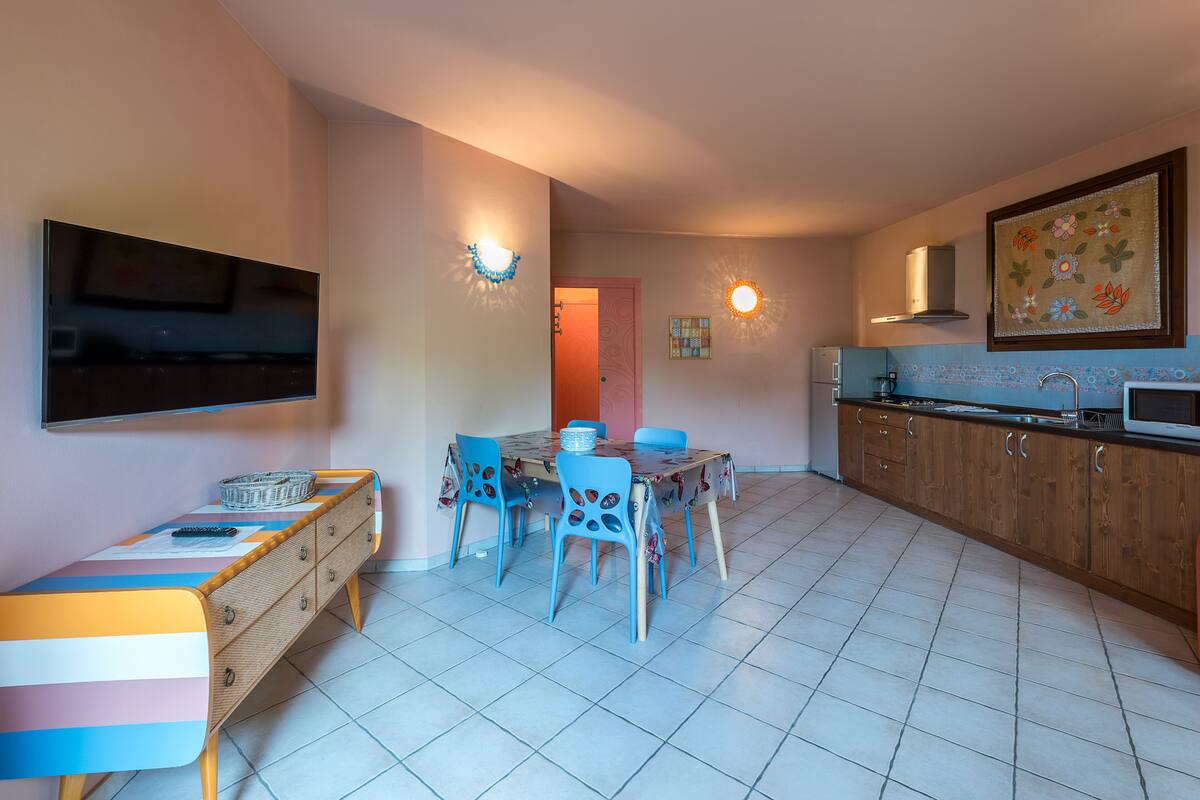 Un comedor con una mesa y sillas, un televisor montado en la pared, y una zona de cocina con armarios y un microondas.