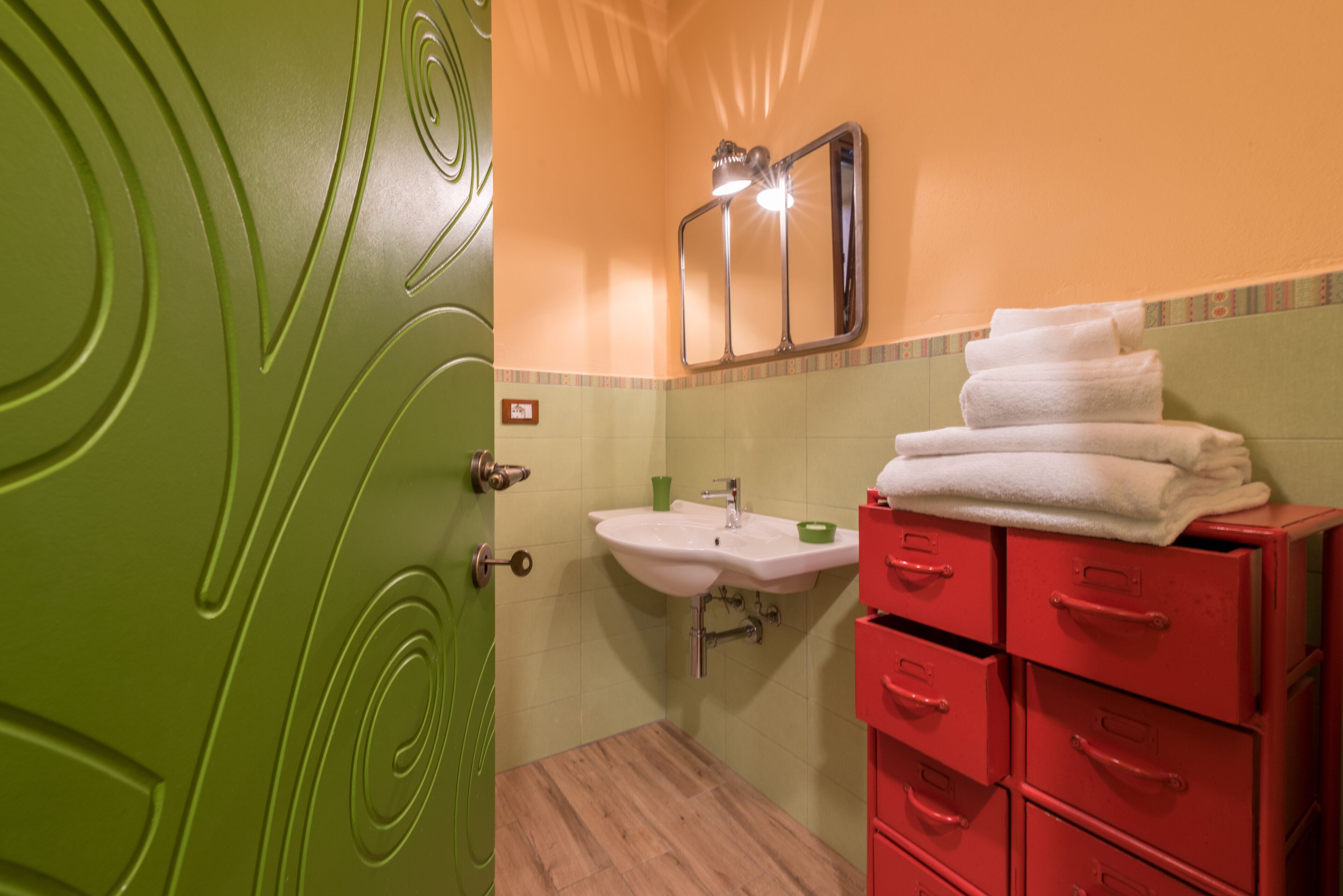 A bathroom with a green door, a sink, a mirror, and a stack of white towels on a red cabinet.
